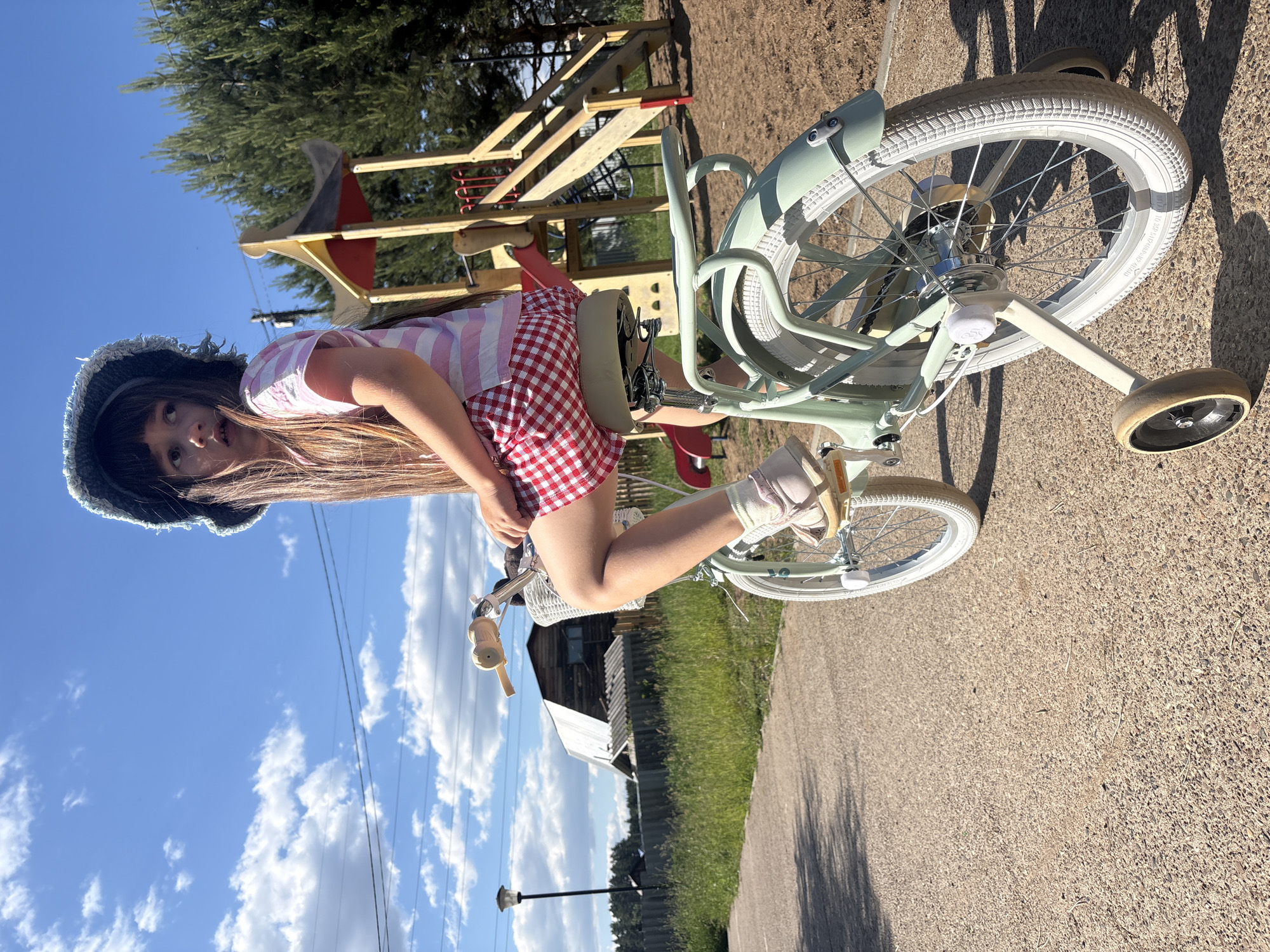 A young girl is positioned on a mint green child's bicycle equipped with white tires, a white front basket, and training wheels, situated on a light-colored asphalt or gravel path. She has long dark hair with bangs, wears a dark, textured bucket hat, a pink and white striped t-shirt, red and white checkered shorts, and light-colored sandals over socks. Her head is turned to her left, with a direct gaze. In the midground, to the left, a black streetlight pole stands before a dark wooden fence and a distinctive light-colored wooden structure with a corrugated roof, possibly a house or barn. Green grass and scattered weeds line the path and the fence. To the right, in the background, a wooden playground structure with a red slide, stairs, and yellow roof accents is visible, surrounded by tall green coniferous trees. The sky is bright blue with scattered white cumulus clouds, and thin power lines traverse the upper left portion of the frame. The scene takes place outdoors in Chaikovsky, Russia, under clear daylight conditions.