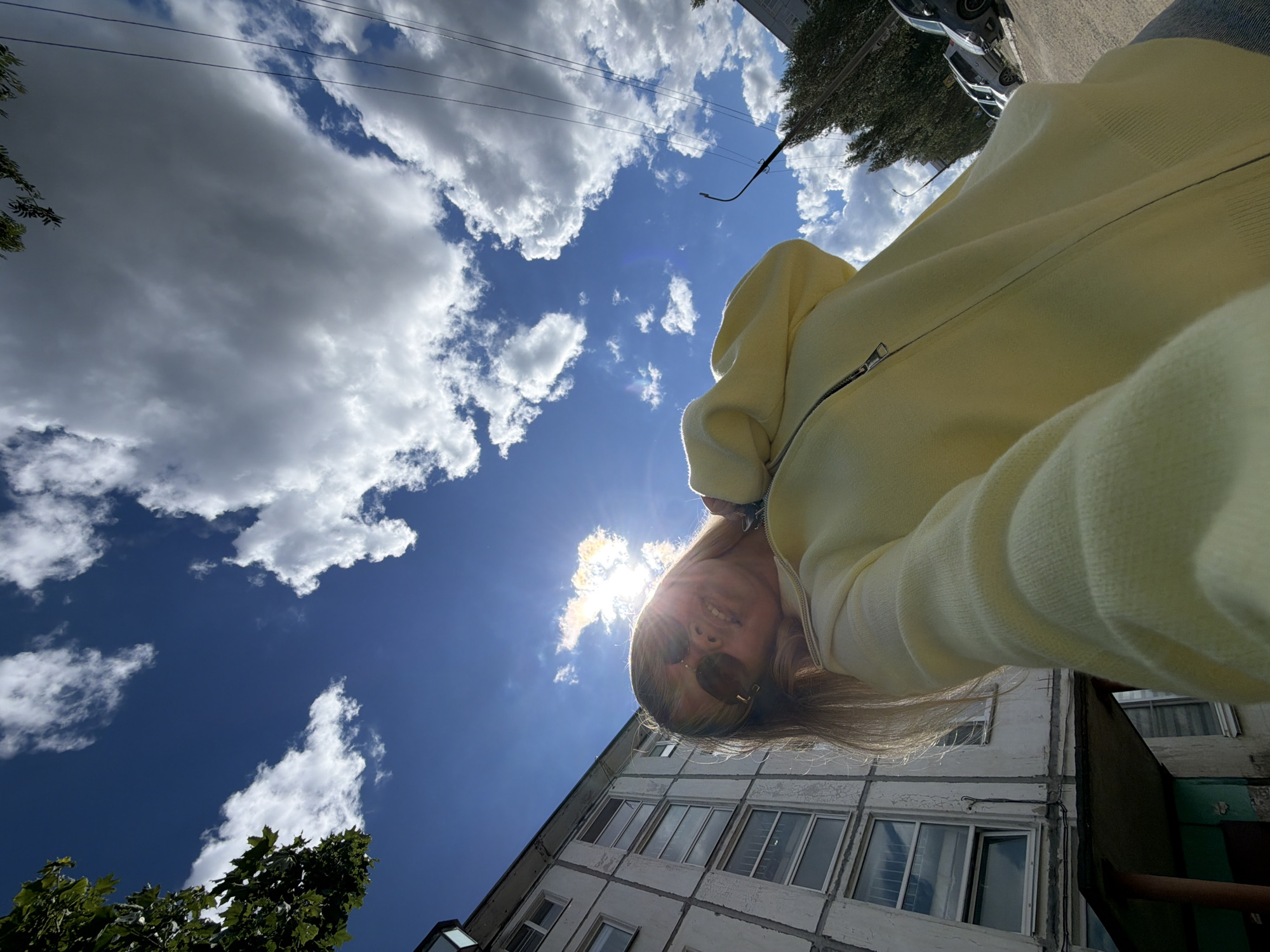 A low-angle shot captures a person wearing a yellow zip-up hoodie and sunglasses, with the sun positioned behind them. The background is composed of a bright blue sky dotted with puffy white clouds. A building with numerous windows is visible in the lower right corner of the frame. Based on the architecture and surrounding environment, the location is likely within the city of Chaikovsky, Russia. Parts of trees and utility lines are also visible in the top corners. The person's face is partially obscured by the sun's glare, suggesting that the photo was taken facing the sun.