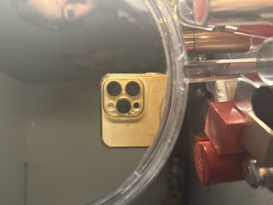 A person’s reflection appears in a round mirror. Within the mirror’s reflection, a gold iPhone with three rear cameras is prominent. The mirror is surrounded by various items, including several lipsticks, some in red packaging, creating a cluttered border around the reflective surface.