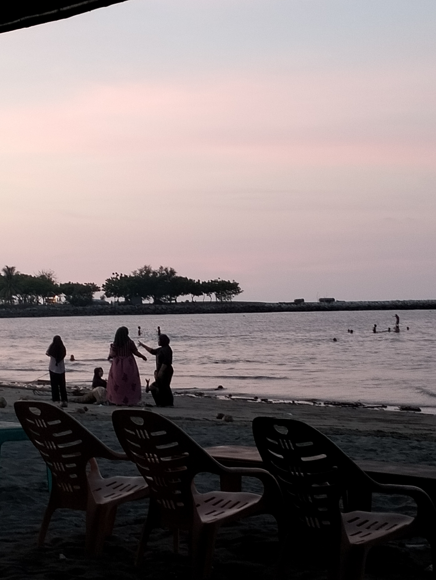 This image captures a serene coastal scene at dusk or dawn, likely in Lhokseumawe, Indonesia. The sky dominates the upper portion of the frame, displaying soft gradients of light pink, purple, and pale blue, indicative of the early or late hours of the day.
In the foreground, three dark, silhouetted plastic chairs face the water, suggesting a spot for relaxation or dining by the shore. The middle ground features a sandy beach where several people are gathered. Their figures are largely silhouetted against the water and the sky. One person wearing a long, light-colored dress stands prominently, interacting with others. To their left, another individual stands facing the water, while a few others are seated or reclining on the sand.
Further out in the calm, reflective water, several people are seen wading or swimming, appearing as small, dark shapes. A distant treeline and a low, dark structure, possibly a breakwater or artificial barrier, define the horizon in the background. The overall impression is one of peaceful recreational activity at a beach as the day transitions. There is no visible text in the image.