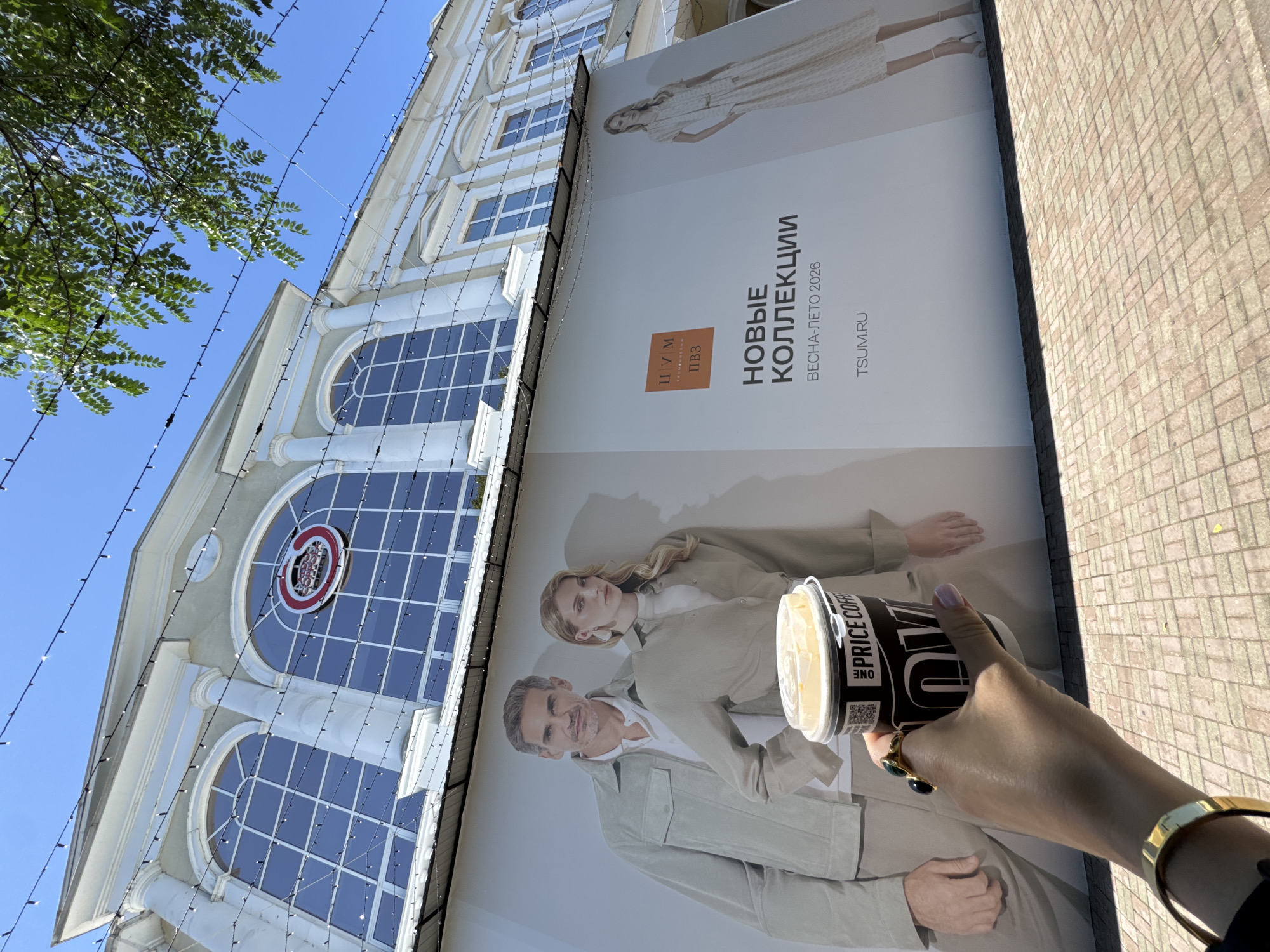 This image captures a street scene in Sochi, Russia, under a clear blue sky. The foreground shows a person's hand, adorned with rings and a bracelet, holding a takeaway coffee cup. The cup is black with white text, featuring "ONE PRICE COFFEE" prominently.
In the background, a large building with ornate architecture is visible, its facade decorated with string lights. A large billboard is plastered on the building, displaying a fashion advertisement. The billboard features two models, a man and a woman, dressed in neutral-toned clothing. Text on the billboard in Russian reads "НОВЫЕ КОЛЛЕКЦИИ" (New Collections) and "ВЕСНА-ЛЕТО 2026" (Spring-Summer 2026), along with "TSUM.RU". A logo with Cyrillic letters "ЦУМ" is also present.
The lighting suggests it is daytime, likely late morning or early afternoon, given the bright sunlight and clear skies, casting shadows on the tiled pavement. The overall mood is casual and urban, with the focus shifting between the person enjoying their coffee and the imposing building adorned with advertisement and lights. The image appears to be taken from a low angle, looking upwards.