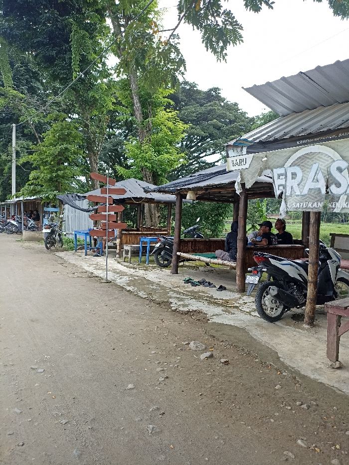 The image depicts a casual roadside scene in Kepung, Indonesia, featuring several open-air huts or stalls serving as gathering spots, likely for food and drink. The foreground shows a dirt or gravel road, with a partially cemented pathway running alongside the huts.
Multiple people are visible within the huts. In the rightmost hut, several men are seated on a bamboo bench, conversing and relaxing. Other individuals are partially visible in the central and leftmost huts. Numerous motorcycles (scooters) are parked near the huts, indicating customer presence. Neatly placed sandals and slippers are visible on the ground beside the rightmost hut, suggesting patrons have removed their footwear before sitting.
A brown, multi-directional wooden signpost with blank pointers stands prominently in the foreground. The huts are constructed with simple bamboo or wooden frames and corrugated metal roofs, providing shade. Lush green trees form a dense backdrop, suggesting a natural or semi-rural setting.
A torn banner or sign on the front of the rightmost hut contains partially visible Indonesian text. Fragments read "BARU" (meaning "New"), "FRA SA," "SATURAN KEN," and "KARENA SENYUMMU SAJA" (meaning "Because of your smile alone").
The scene appears to take place during the daytime under diffused light, possibly on an overcast day, conveying a relaxed and informal atmosphere.