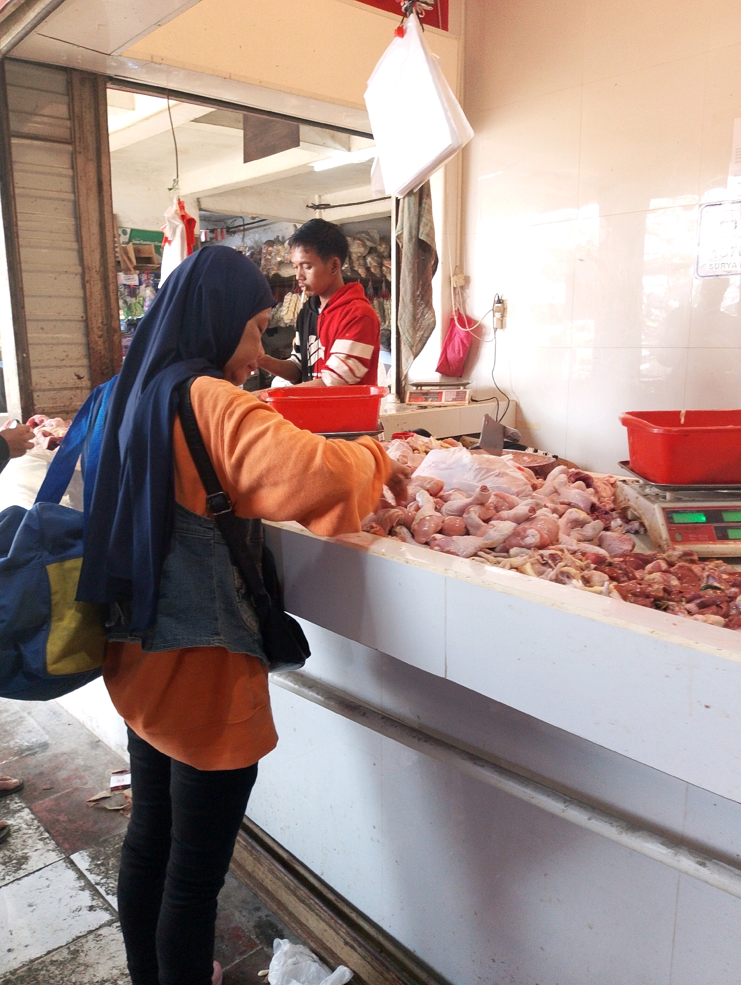 A woman wearing a navy hijab, an orange long-sleeved shirt, and black leggings stands at a butcher stall in a market in Lembang, Indonesia. She is reaching for a large quantity of raw chicken displayed on a white counter. A blue and yellow bag is slung over her left shoulder, and a black bag is on her right shoulder. Behind her, a man in a red and white shirt is visible. The stall is filled with raw chicken parts, some in plastic bags. Scales are visible on the counter, and red plastic containers are present. The environment appears to be an indoor market with tiled walls and wooden stall structures.