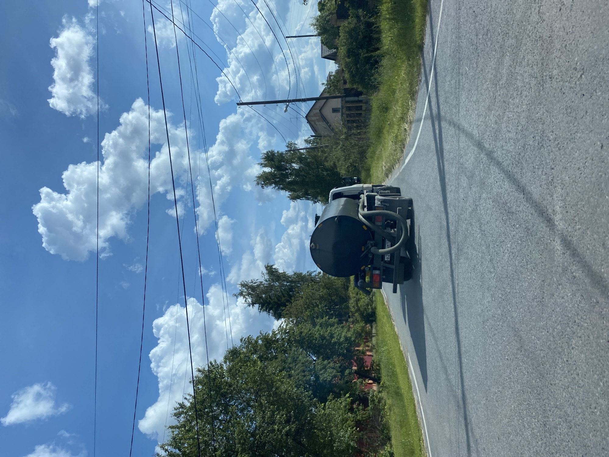 An empty black tanker truck drives down a paved road on a clear day in Tyumen, Russia. The truck has a large cylindrical tank mounted on its chassis. The road is bordered by green grass and trees on both sides. Houses with red and brown roofs are visible in the distance, nestled among the trees. Overhead, power lines crisscross the bright blue sky, which is dotted with fluffy white clouds. The scene is serene and suggests a rural or suburban setting within Tyumen.