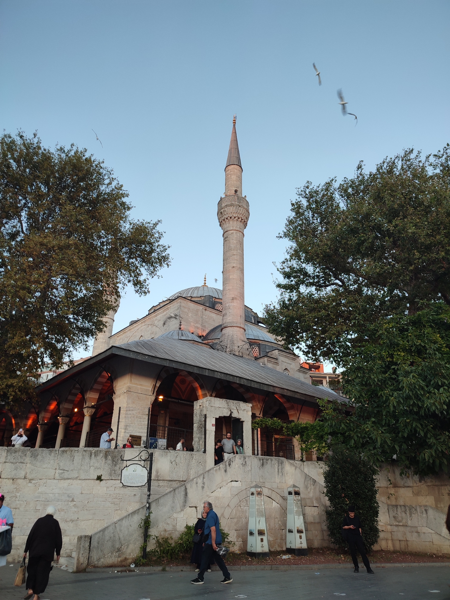 This image shows the Mihrimah Sultan Mosque complex in Üsküdar, Turkey, viewed from street level. The mosque features a prominent minaret and a series of domes, constructed with light-colored stone. The foreground depicts a stone retaining wall with stairs leading up to the mosque's elevated courtyard. Several people are visible in the scene: a person in a black outfit walks away from the camera in the lower left, while others are seen on the stairs and in the courtyard area. Two white, obelisk-shaped informational displays are positioned on the street in front of the wall. Large trees with dense green foliage flank the mosque. The sky above is a clear, pale blue with two birds in flight. The lighting suggests it is late afternoon or early evening, with warm light illuminating the side of the minaret and the arched portico of the mosque.