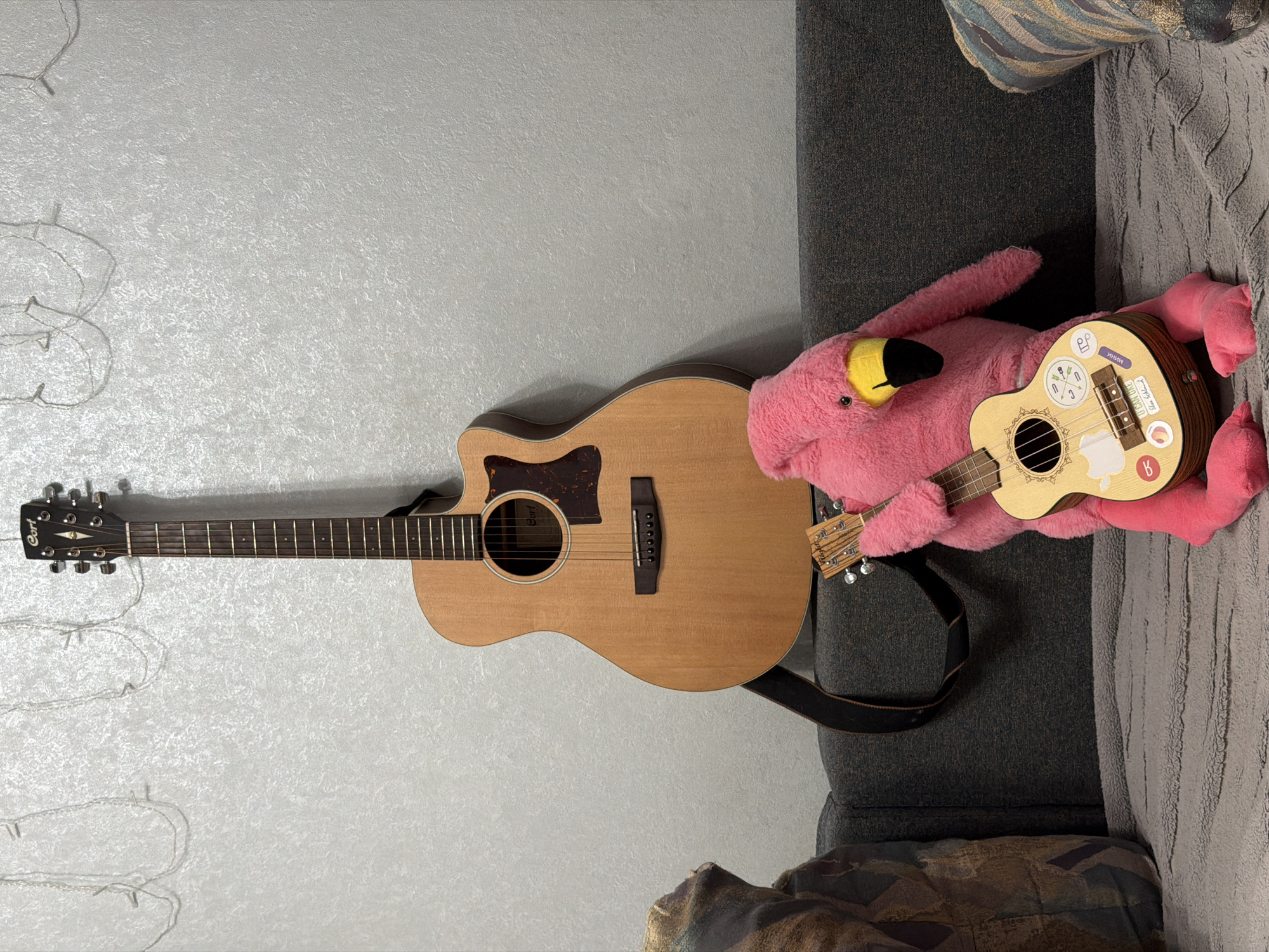 A large, light brown acoustic guitar stands upright behind a stuffed pink flamingo wearing a smaller ukulele. The flamingo is seated on a grey couch, positioned to the right of the guitar. The background consists of a textured white wall with decorative fairy lights draped across the top. The scene appears to be indoors, likely in a residential setting. There is no visible indication of the city of Chaikovsky, Russia.