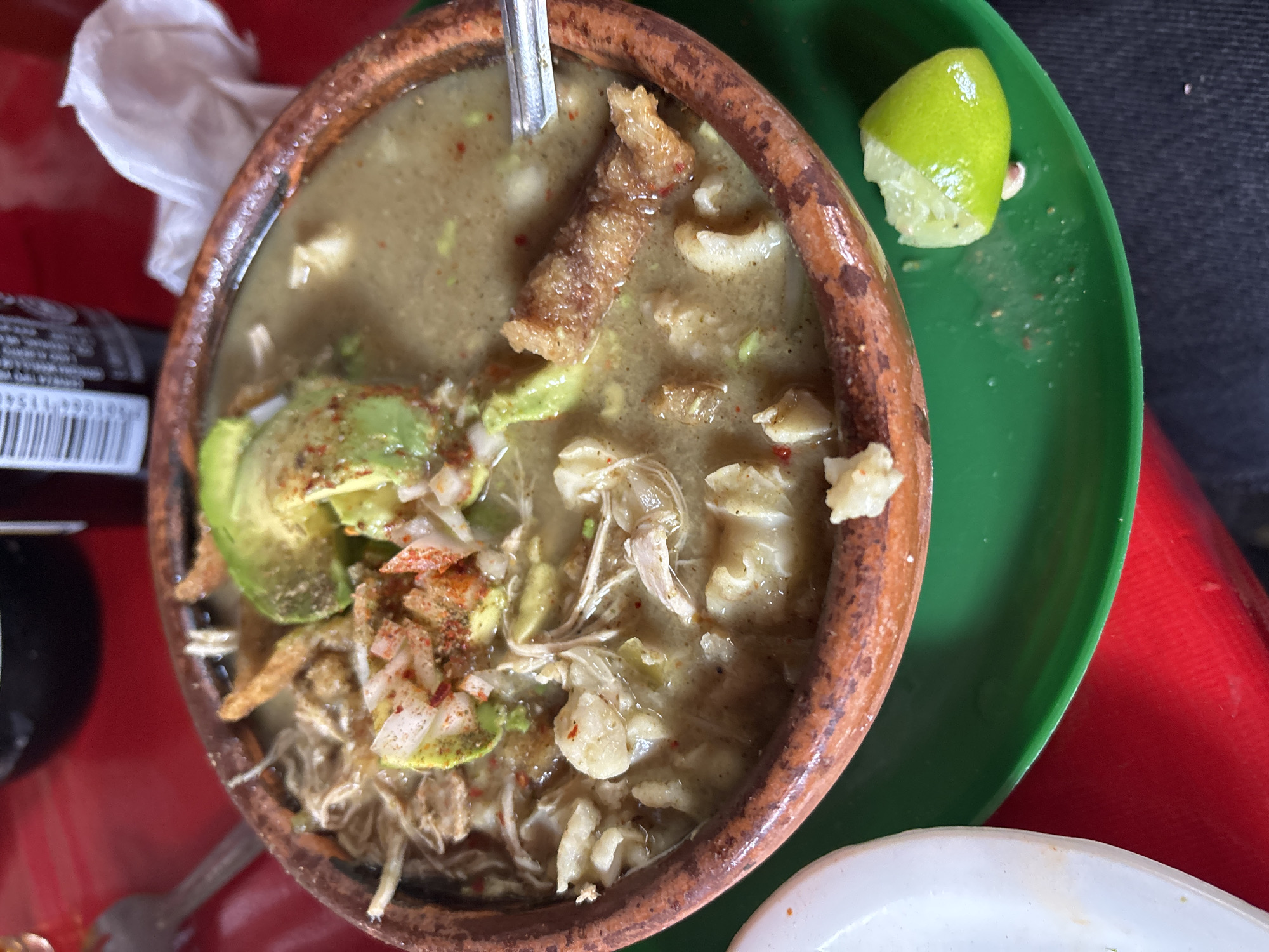 A bowl of green pozole sits on a red table in Taxco, Mexico. The pozole, served in a brown clay bowl, is garnished with shredded chicken, hominy, avocado slices, chicharron, onion, and a lime wedge. A green plastic plate lies underneath the bowl. A spoon is visible inside the bowl. A bottle and a white napkin rest on the table next to the pozole.