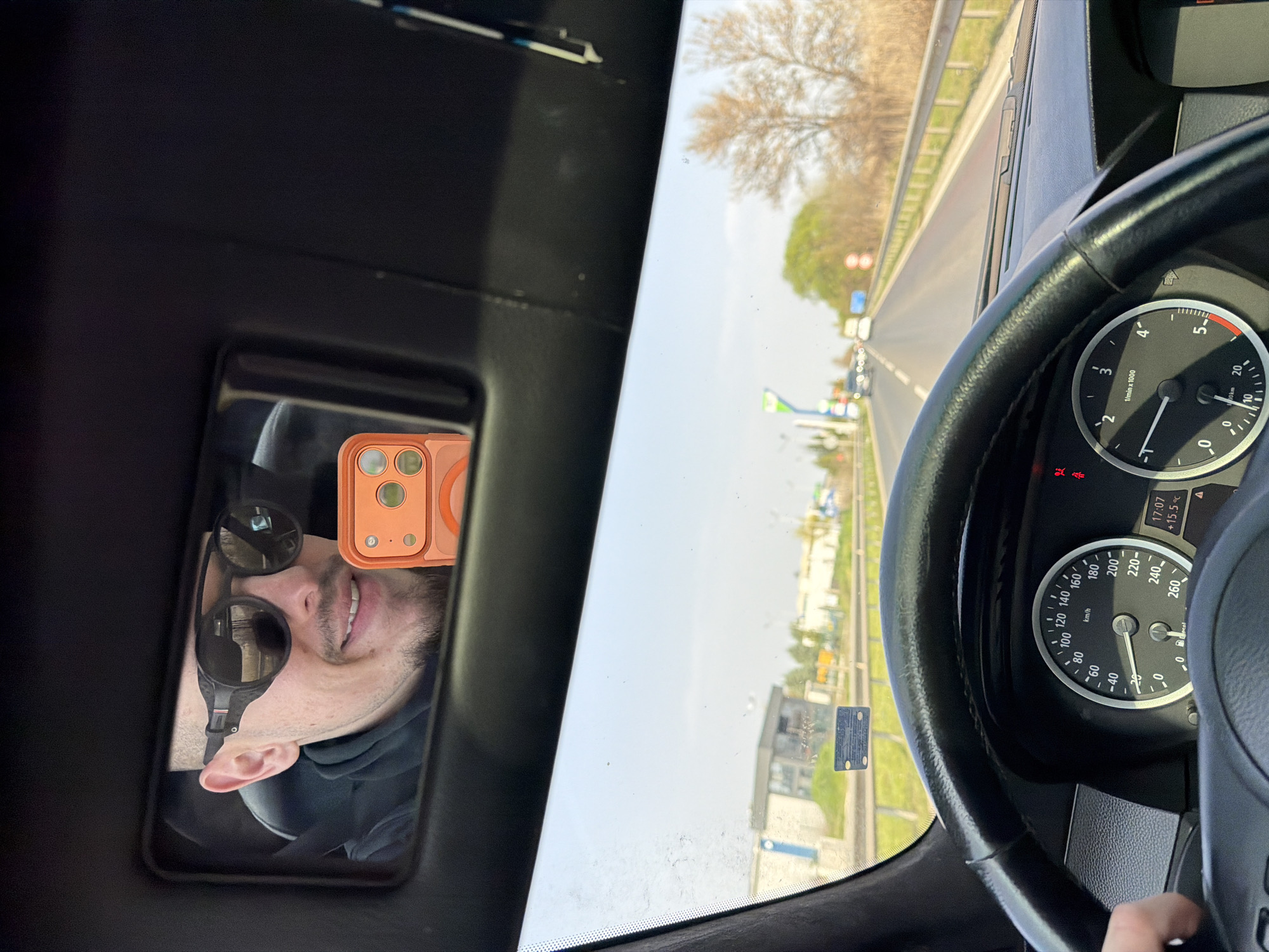 The image shows the interior of a car, presumably in motion. A man's reflection is visible in a mirror; he is wearing sunglasses and seems to be holding a phone. Through the windshield, a road is seen with trees and buildings in the background. The car's dashboard displays a speedometer reading approximately 100 km/h and a tachometer around 3.5 x 1000 RPM. The location is likely Plovdiv, Bulgaria.