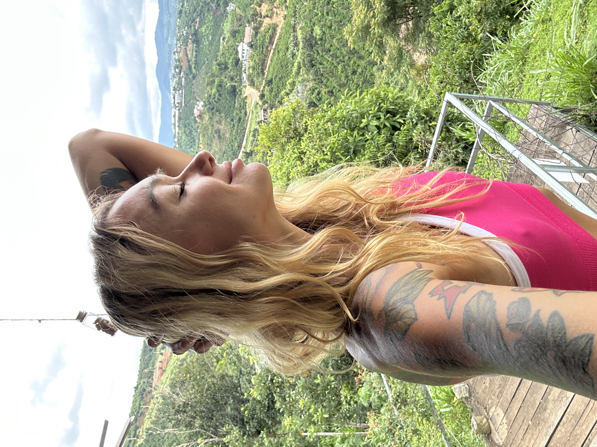 The image shows a woman with blonde hair lying down with her head propped up on her arm. She is wearing a bright pink top with white trim. Her arm, visible in the foreground, is adorned with tattoos. The background features a lush, green, mountainous landscape with some buildings visible in the distance. The sky is overcast with clouds. The scene appears to be outdoors, possibly on a balcony or terrace, with wooden planks visible in the foreground and a metal railing to the right. The orientation of the image suggests it was taken in a vertical position. The overall mood is relaxed and contemplative, with the woman's eyes closed and her head tilted back. The location is identified as Thôn 3, Vietnam.