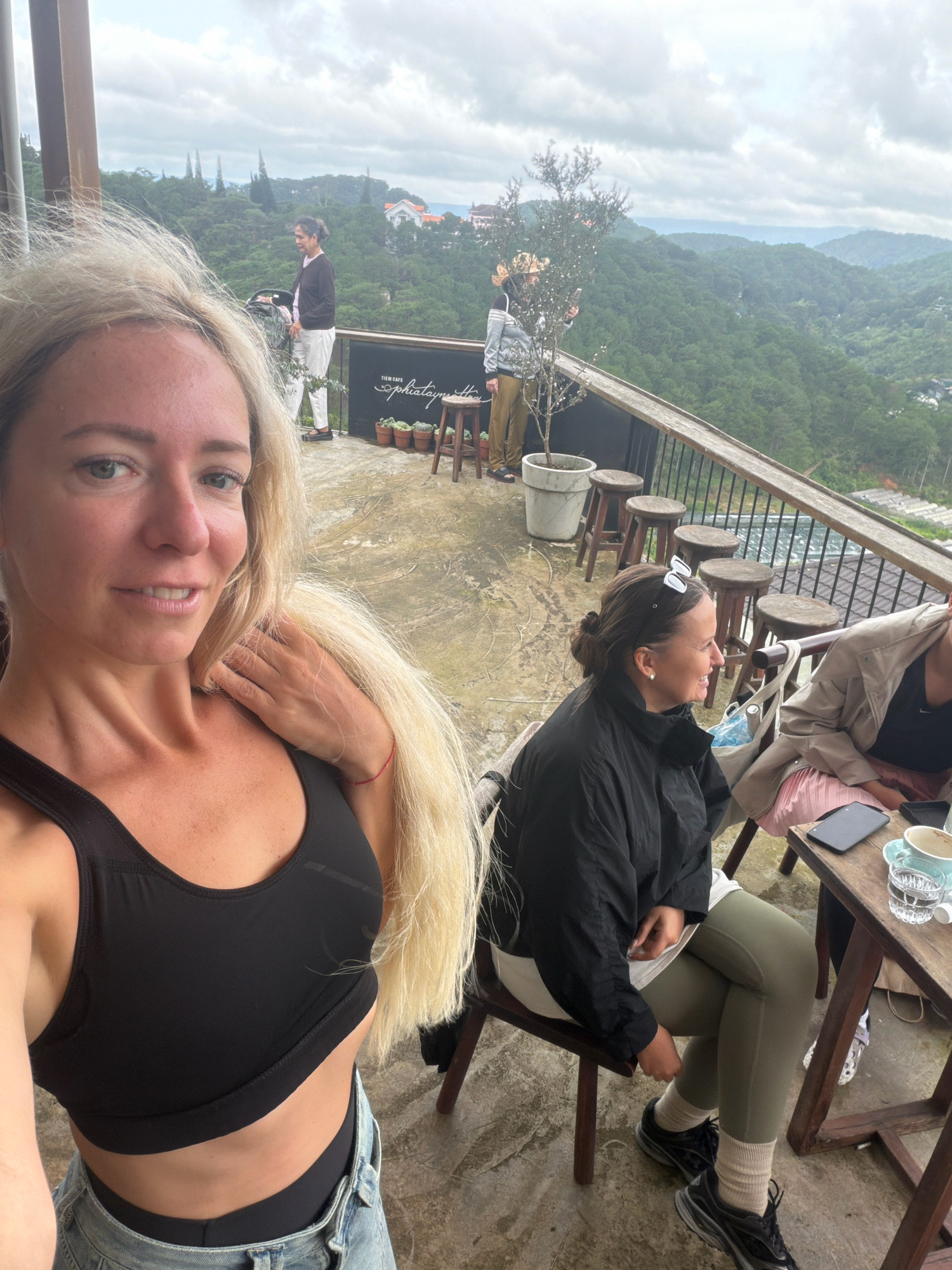 The image shows four women on an outdoor terrace with a view of lush green hills in Da Lat, Vietnam. In the foreground, a woman with blonde hair is taking a selfie. She is wearing a black sports bra and denim jeans. To her right, another woman with dark hair is sitting on a chair, wearing a black jacket and olive-green leggings. Next to her, a third woman, partially visible, is wearing a light-colored jacket over a pink skirt. A fourth woman is standing in the background, wearing a dark top and white pants, and appears to be holding a phone. The terrace has a rustic wooden railing and several wooden stools. In the background, the landscape is characterized by rolling hills covered in dense vegetation and some scattered buildings. The sky is overcast. The sign "THEM CAFE Sophiatime" is visible on a dark panel behind the standing woman.