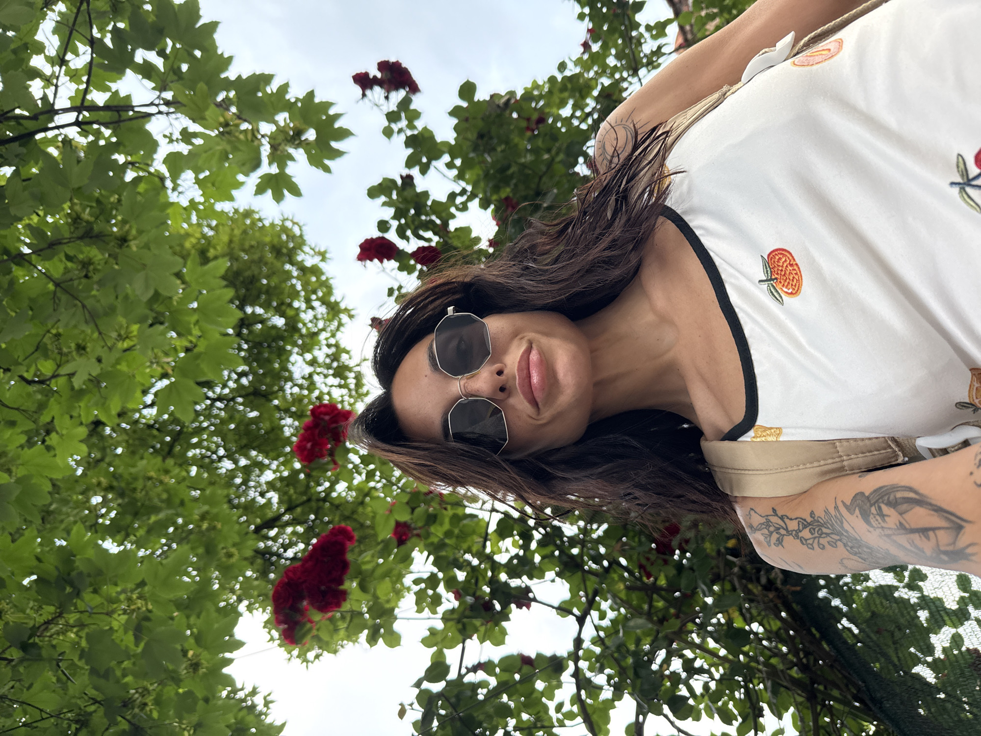 The image displays a low-angle, eye-level portrait of a young adult female, positioned outdoors in the city of Pleven, Bulgaria. The subject has long, dark, wavy hair that appears slightly damp. She wears dark-lensed, octagonal-framed sunglasses, obscuring her eyes. Prominent freckles are visible across her nose and upper cheeks. Her lips are full, glossy, and slightly parted. She is attired in a white tank top featuring a black trim along the neckline and embroidered fruit motifs, including a distinct orange or tangerine on the upper chest. A light-colored strap, consistent with a backpack or bag, is visible over her right shoulder. Tattoos are present on both arms: a detailed floral or vine-like design extending up her left forearm, a stylized female face on the inner left forearm, and a circular motif on her upper right arm. The subject's head is tilted slightly back, and her gaze is directed downward toward the camera with a subtle smirk. The background is dominated by dense green foliage, consistent with trees featuring broad leaves, and numerous clusters of vibrant red roses, suggesting a flowering bush or vine. An overcast, bright sky is visible through gaps in the foliage, primarily in the upper left quadrant. The scene depicts an outdoor setting beneath a natural canopy.