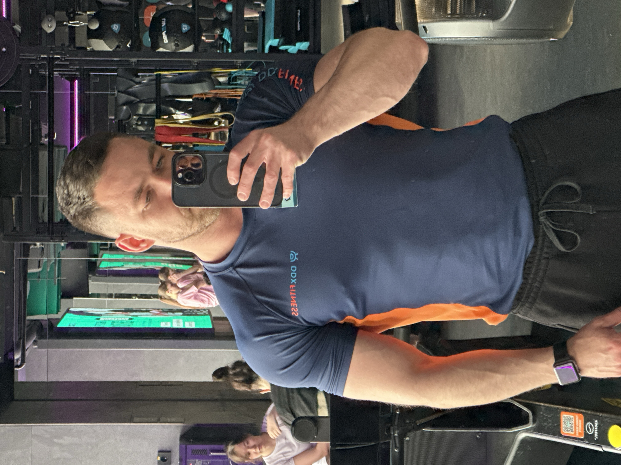 A man is taking a selfie in front of a mirror. He is wearing a navy blue and orange shirt, with the words "DDX FITNESS" printed on it, and black shorts. He is holding a smartphone in his left hand. The mirror reflects a gym with various equipment, including weights and exercise machines. In the background, two women are visible near some gym equipment. The city is not discernible from the image.