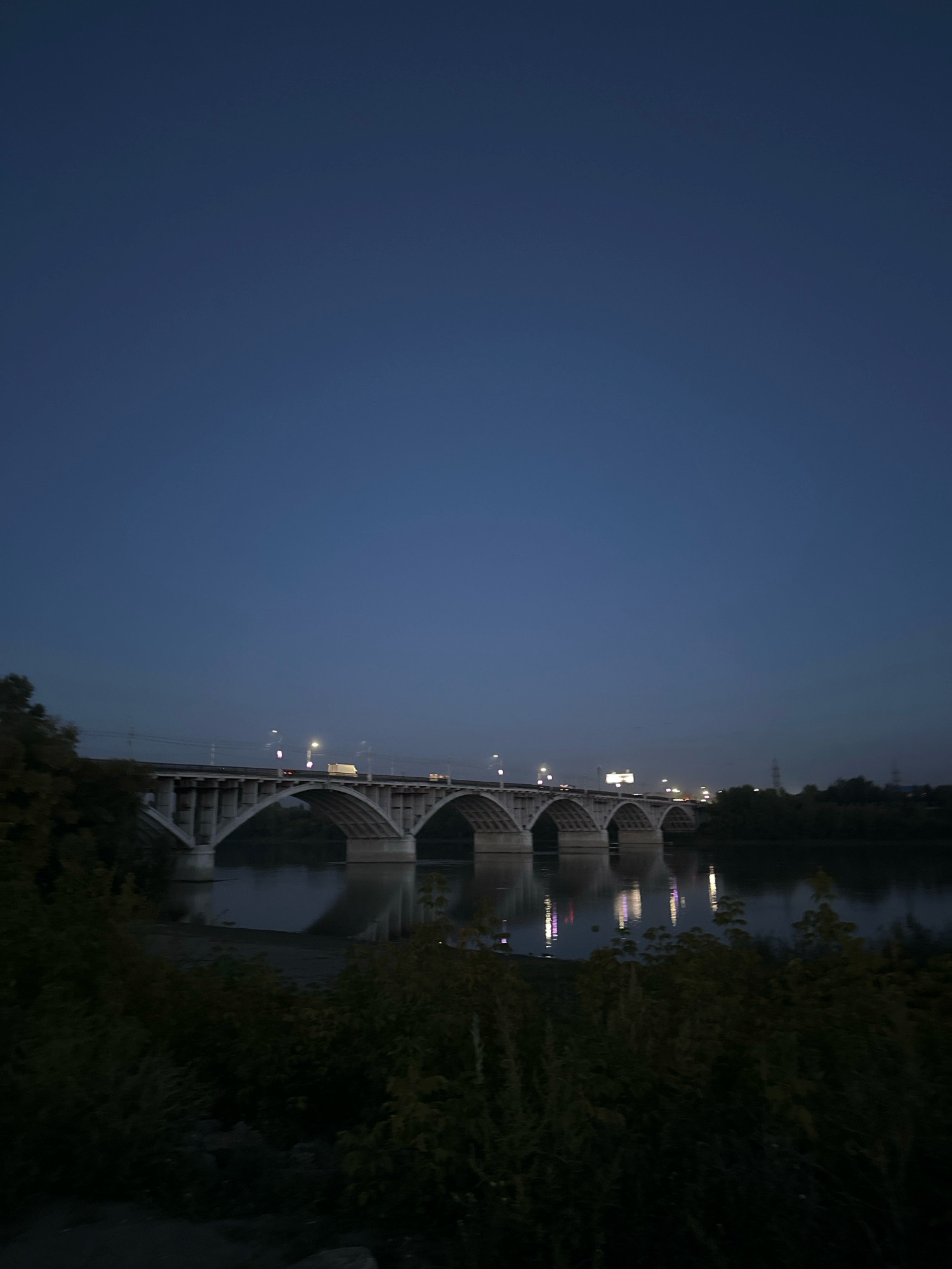 The image shows a bridge spanning a body of water at dusk. The sky is a deep blue, with the lights of cars and streetlamps visible on the bridge. The bridge itself is made of concrete with multiple arches, and its reflection is visible in the calm water below. Trees and foliage line the foreground, casting the lower portion of the image in shadow. The scene appears to be a city bridge, possibly in Biysk, Russia, given the context. There are no visible people in the image. The overall atmosphere is tranquil and somewhat serene, characteristic of twilight in an urban environment.