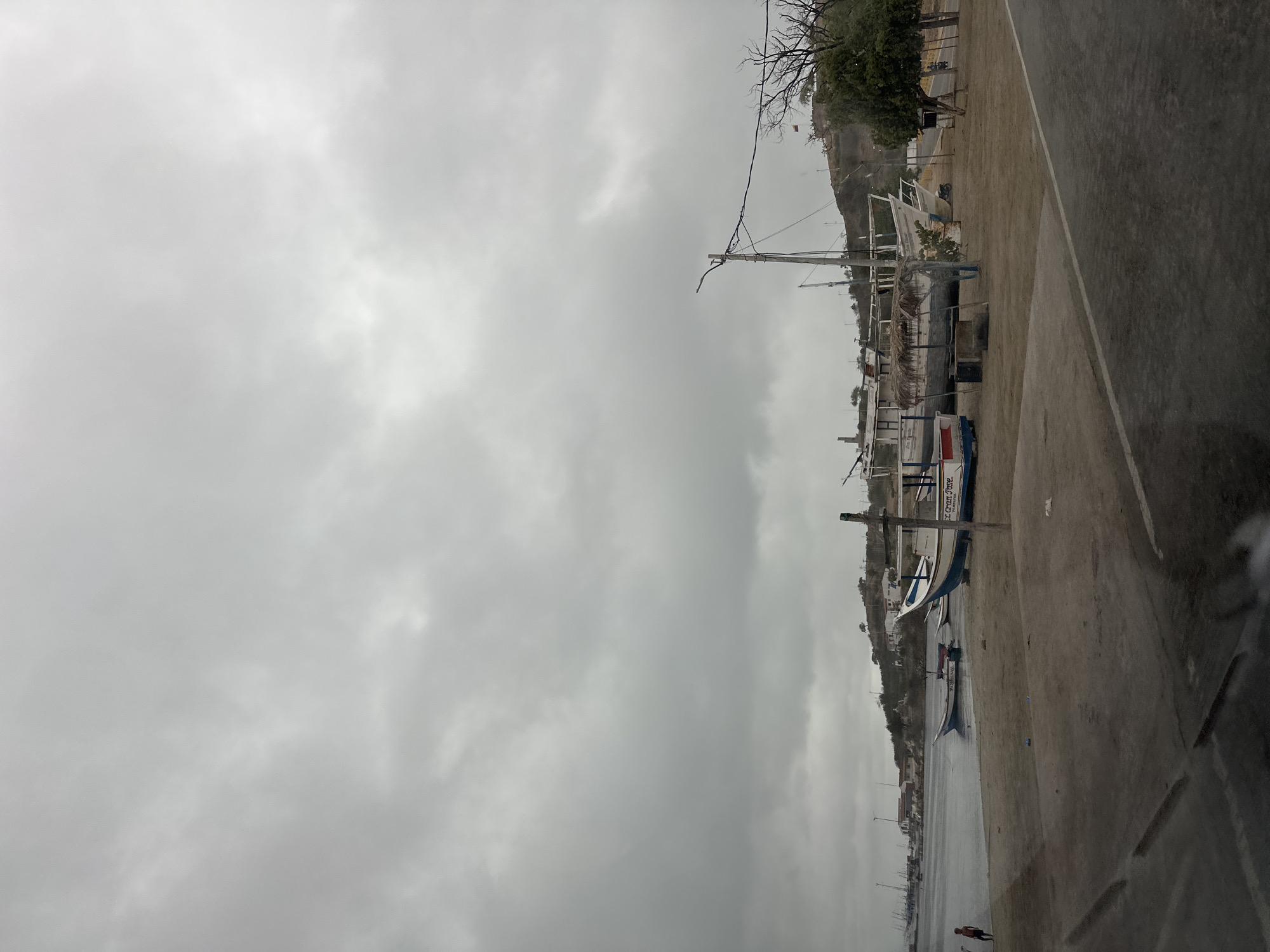 The image depicts a cloudy day scene in Juan Griego, Venezuela, specifically near the shoreline. In the foreground, a concrete area with visible tire marks suggests a road or parking lot. Beyond this, a sandy beach is populated with several boats. One boat, prominently featured on the sand, is a larger vessel with a thatched roof structure and appears to be undergoing some form of work or maintenance. Smaller fishing boats are also visible, some pulled up onto the sand, and one smaller boat is in the shallow water.
In the background, the town of Juan Griego is visible on a gentle slope. White-washed buildings with red roofs are scattered across the hillside. The water's edge is lined with more boats, appearing to be docked or moored. A person is standing near the water's edge on the left side of the image. The sky is overcast with heavy grey clouds, casting a subdued light over the entire scene. A tall utility pole with wires stretches across the middle ground, and bare trees are present on the right side of the image, suggesting a coastal environment. A flag is visible on a pole on the hill in the distance.