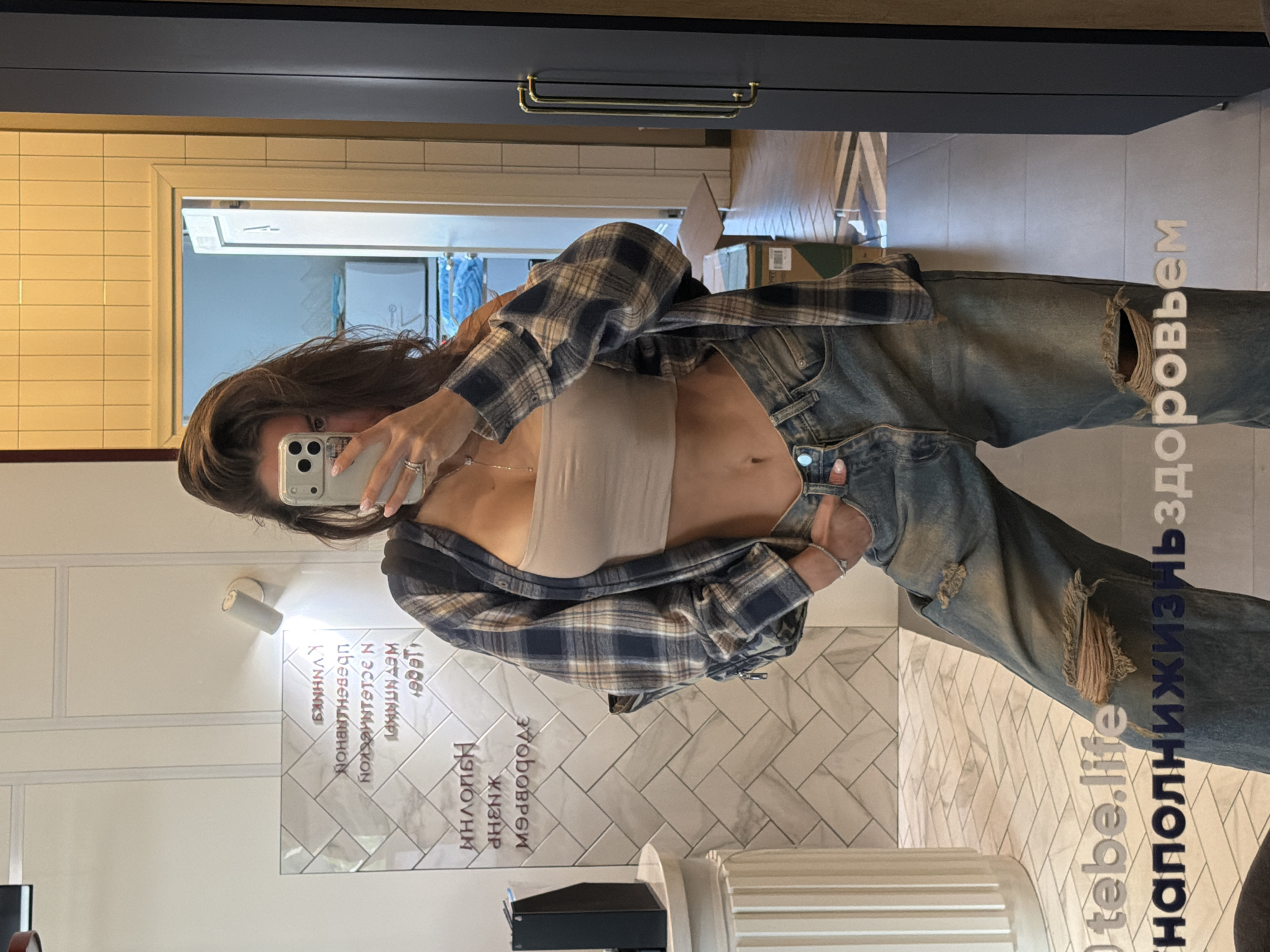 A woman in a casual outfit (tan tube top, flannel shirt, ripped jeans) is taking a selfie in a bathroom. The bathroom has white and gray tiled walls and a dark blue cabinet with gold handles. Text in Russian is written on the mirror: "Наполни Жизнь Здоровьем tebe.life." The woman is standing in front of a white, column-like structure. The exact location within Krasnoyarsk, Russia cannot be determined from the provided image.