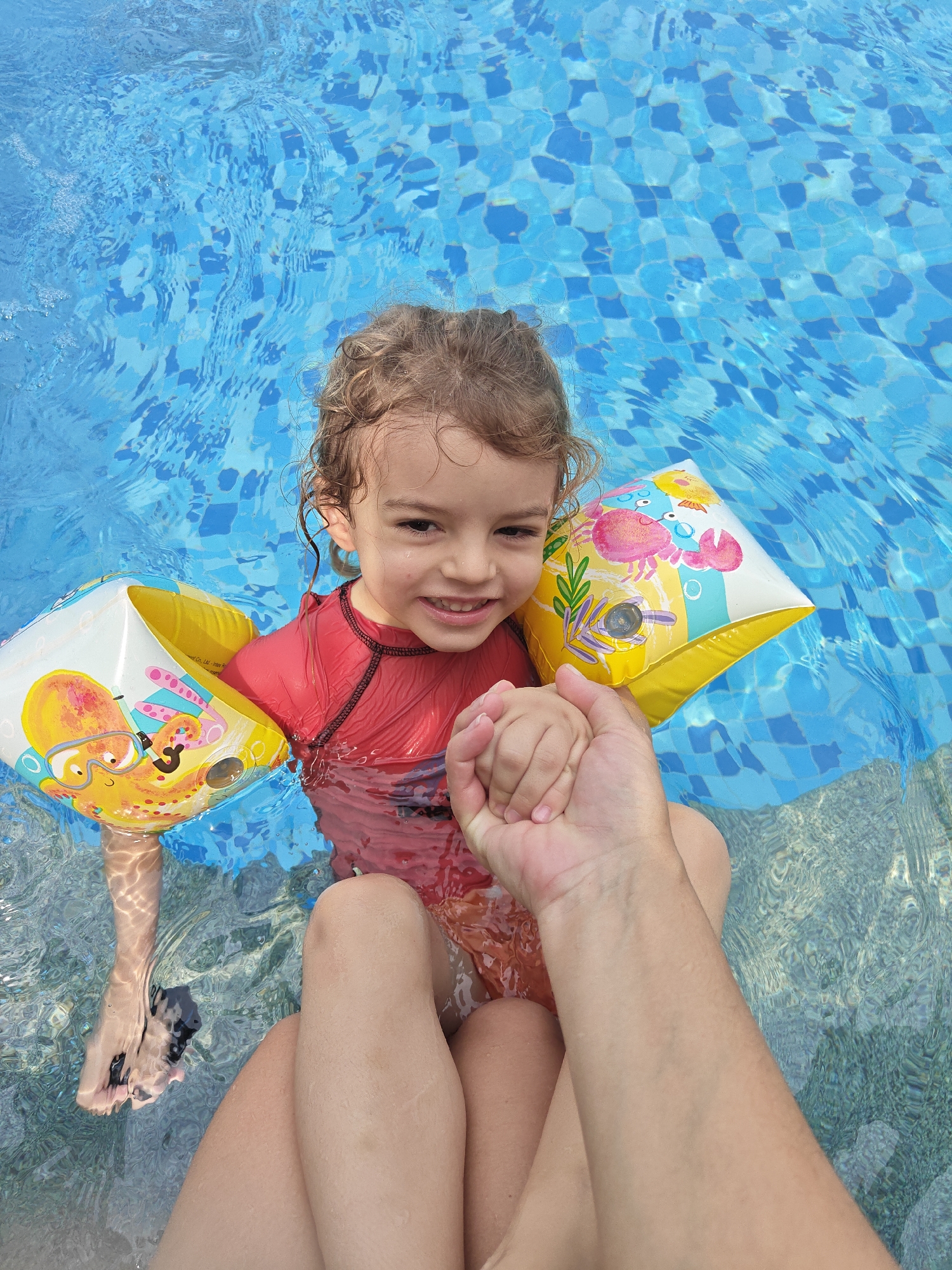 A young child with curly light brown hair, wearing a red rash guard and yellow and white inflatable arm floaties, is sitting in a swimming pool. The pool has blue and white mosaic tiles. The child is holding hands with someone, presumably an adult, whose arm is visible. The setting is likely recreational, such as a swimming pool at a private residence, hotel, or water park. Without additional information, it is not possible to confirm that the location is Mumbai, India.