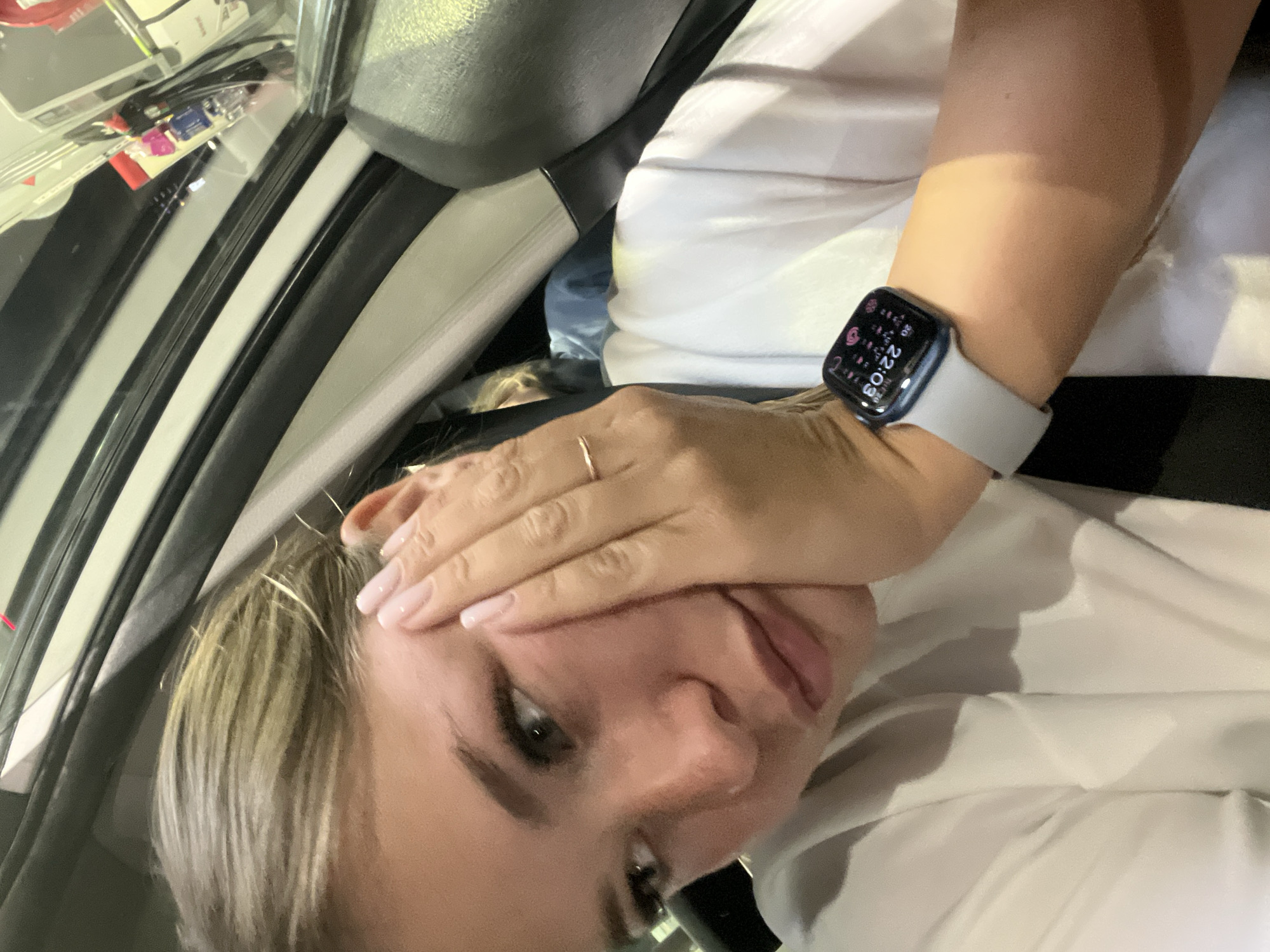 A young woman is captured in a close-up, selfie-style photograph, taken inside what appears to be a car in Budva, Montenegro. She has blonde hair and her head is tilted to the side, looking towards the camera with a slight smile. Her right hand is partially covering her forehead, with her fingers splayed. A thin, gold-colored ring is visible on her right ring finger. She is wearing a white top or t-shirt.
On her left wrist, she wears a modern Apple Watch with a light gray band and a dark screen. The time clearly displayed on the watch face is "22:03," indicating it is 10:03 PM, consistent with the dim, possibly artificial lighting inside the vehicle. Various small app icons and other numerical data are also visible around the time on the watch face, though not clearly legible.
The background shows elements of a car interior, including a seatbelt shoulder strap, the car's door frame or window frame, and what appears to be a dashboard area with some objects like cables and small items visible through the glass. The angle suggests she is a passenger, possibly in the front seat. The overall mood conveyed is casual, perhaps a bit playful or tired, given the time of night.