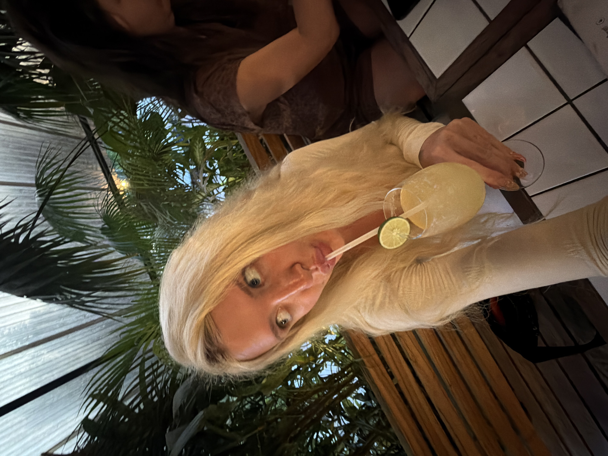A young woman with long blonde hair and blue eyes is taking a selfie while holding a margarita with a lime wedge and a straw. She is wearing a cream-colored, long-sleeved shirt. To her right, another woman with dark hair is seated, wearing a brown patterned top and shorts. They are at an outdoor restaurant with wooden benches and tropical plants visible in the background. The table they are sitting at has a tiled surface. The overall lighting suggests it is evening. The location is identified as North Kuta, Indonesia.