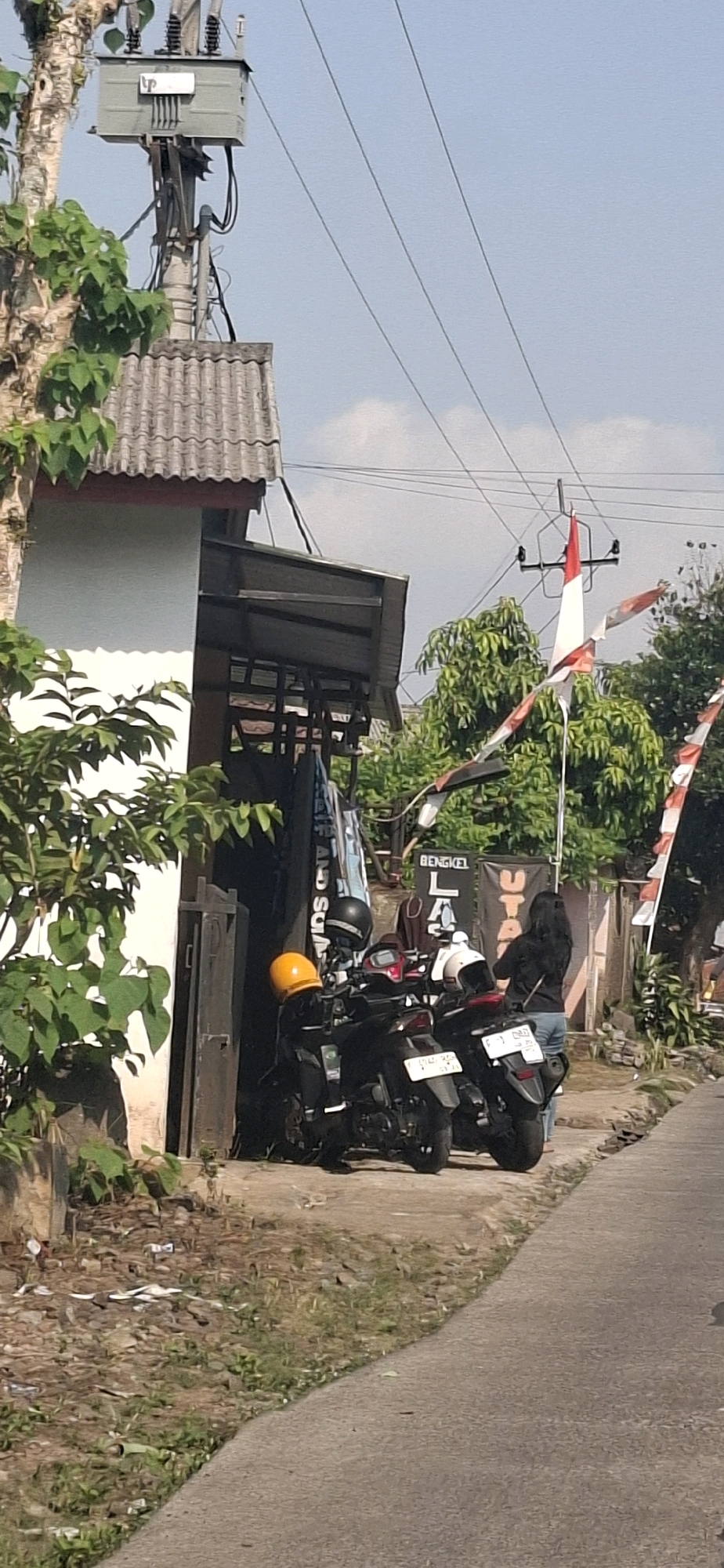 The image depicts a scene on a street in Cibinong Hilir, Indonesia, on a clear day. In the foreground, a gray paved road curves to the right. Adjacent to the road is a patch of dry, uneven ground with scattered debris and sparse vegetation.
Behind this embankment, a structure with a corrugated gray roof is visible, part of a building with a white exterior wall and a brown-trimmed eave. A metal awning extends from the building. Several motorcycles are parked under this awning. One motorcycle in the foreground has a bright yellow helmet on its handlebars. Two other motorcycles are partially visible behind it, and another has a white helmet.
A person, appearing to be a woman with long black hair wearing a black top and blue jeans, stands to the right of the motorcycles, facing away from the camera. Behind her and the motorcycles, several banners are displayed. One banner reads "BENGKEL", suggesting a workshop or repair shop. Indonesian flags, red and white, are strung across the scene, fluttering in the breeze.
To the left of the building, a tree with light-colored bark and green leaves is present. Overhead, electrical wires are strung across the sky, supported by utility poles and a transformer box mounted on one of the poles. The sky is blue with a few scattered white clouds. The overall impression is of a residential or commercial street scene in a tropical environment.