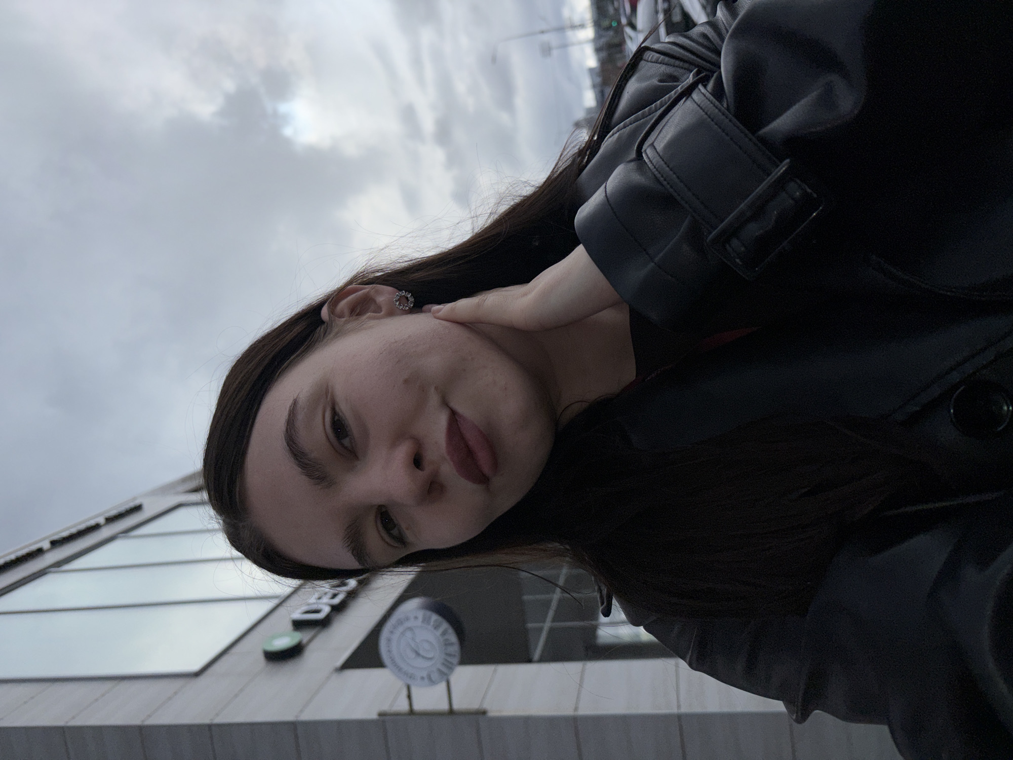 A young woman is depicted in a close-up, upward-angled outdoor portrait, oriented vertically but appearing tilted to her right, suggesting the photo was taken with the camera rotated. She has long, dark hair, fair skin with some visible blemishes on her cheek, and dark, well-defined eyebrows. Her lips are slightly parted, accented with a dark pink or mauve lipstick, and she offers a subtle, relaxed expression. A decorative, possibly silver-toned earring is visible in her left ear, and her left hand is gently resting against her jawline or neck.
She is wearing a black leather-look jacket featuring a collar, visible stitching, and a buckle detail on the sleeve. The attire suggests a cool or temperate weather condition.
The background shows an urban environment with a modern building featuring light-colored facade elements and large, reflective glass windows. A sign on the building partially displays the letters "DEC". Below it, a round, white sign with an indistinct emblem is also visible. The sky is uniformly overcast, indicating a cloudy daytime setting, with a brighter patch of light attempting to break through. The woman appears to be posing for the photograph, potentially in Perm, Russia, given the location context.