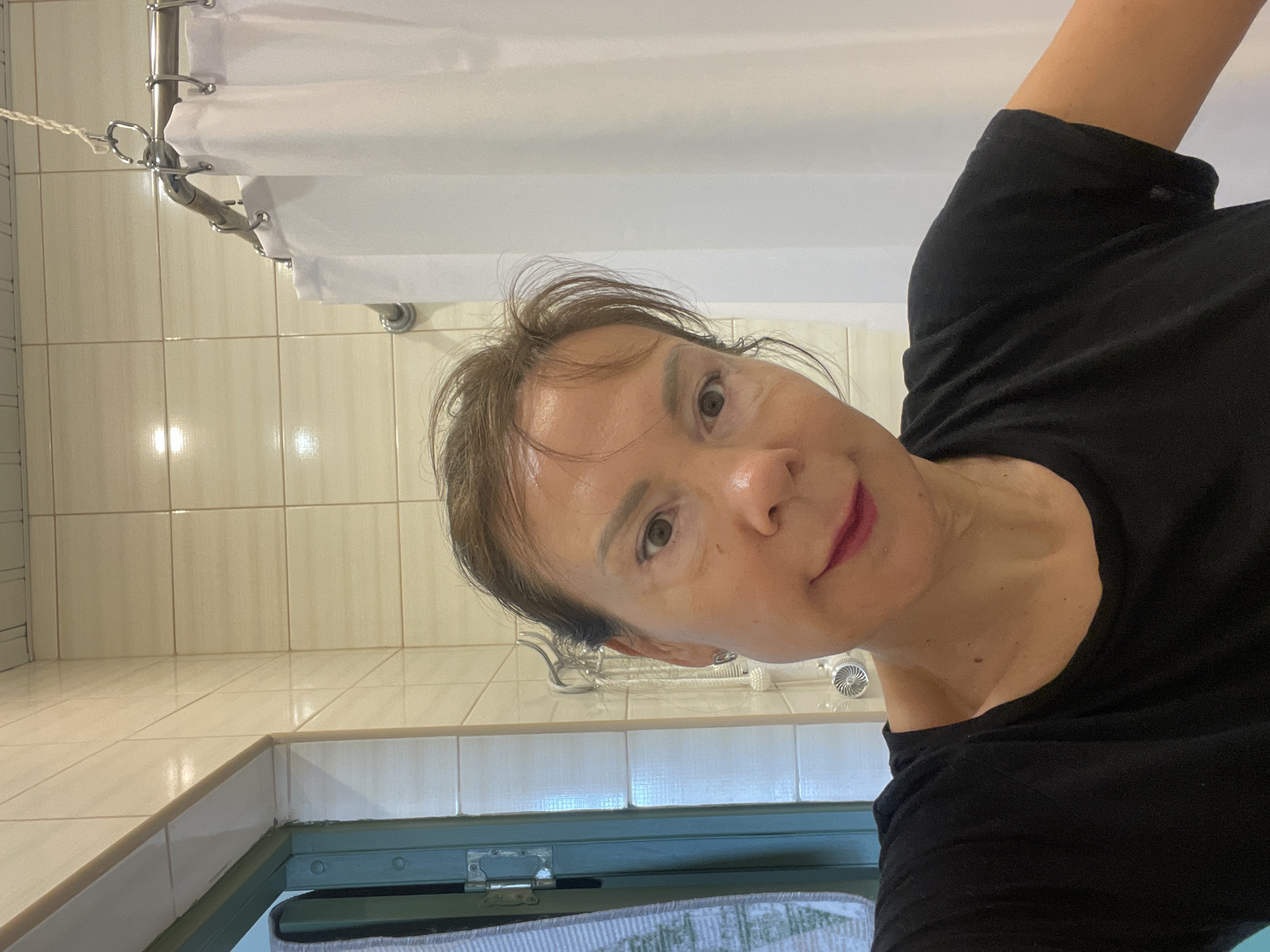 An overhead, medium shot captures a woman in her 50s with medium brown hair and fair skin, wearing a black t-shirt. She is positioned in a bathroom, indicated by the beige tiled walls, a white shower curtain, and a green-framed doorway with a grey and white striped towel hanging from it. The lighting is bright, likely from overhead bathroom lights, creating reflections on her forehead and nose. The woman is looking directly at the camera with a neutral expression.
