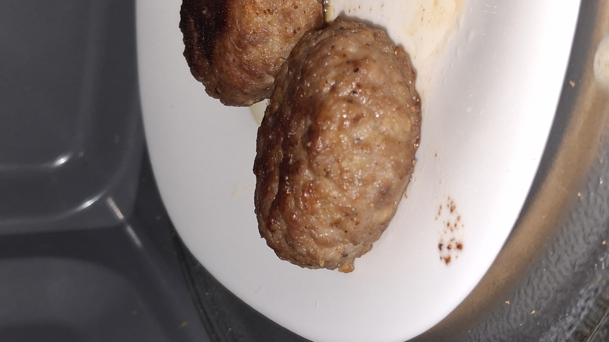 Two meatballs are placed on a white plate. The plate sits on a gray surface with a textured edge. The location is unknown.