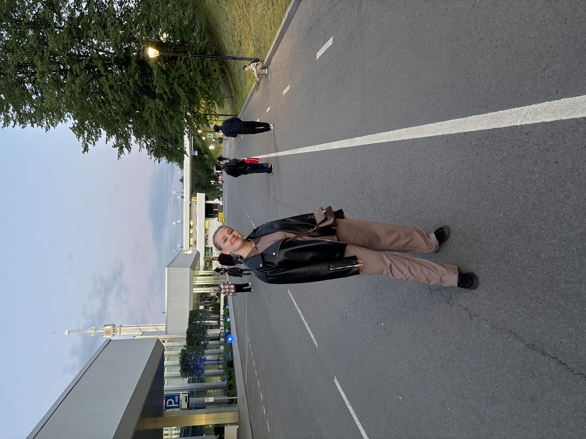 A young woman stands smiling on an asphalt road. She wears a black leather jacket, brown pants, and black shoes. Behind her, other pedestrians walk on the road near a modern building complex with glass facades and trees. A white painted line runs along the road. In the background, a tower resembling a communications or observation tower rises into the sky, suggesting a landmark or significant structure. The image depicts a scene in what appears to be a public space, with the building architecture and tower potentially indicating the location to be in the city of Rusanovo, Russia.
