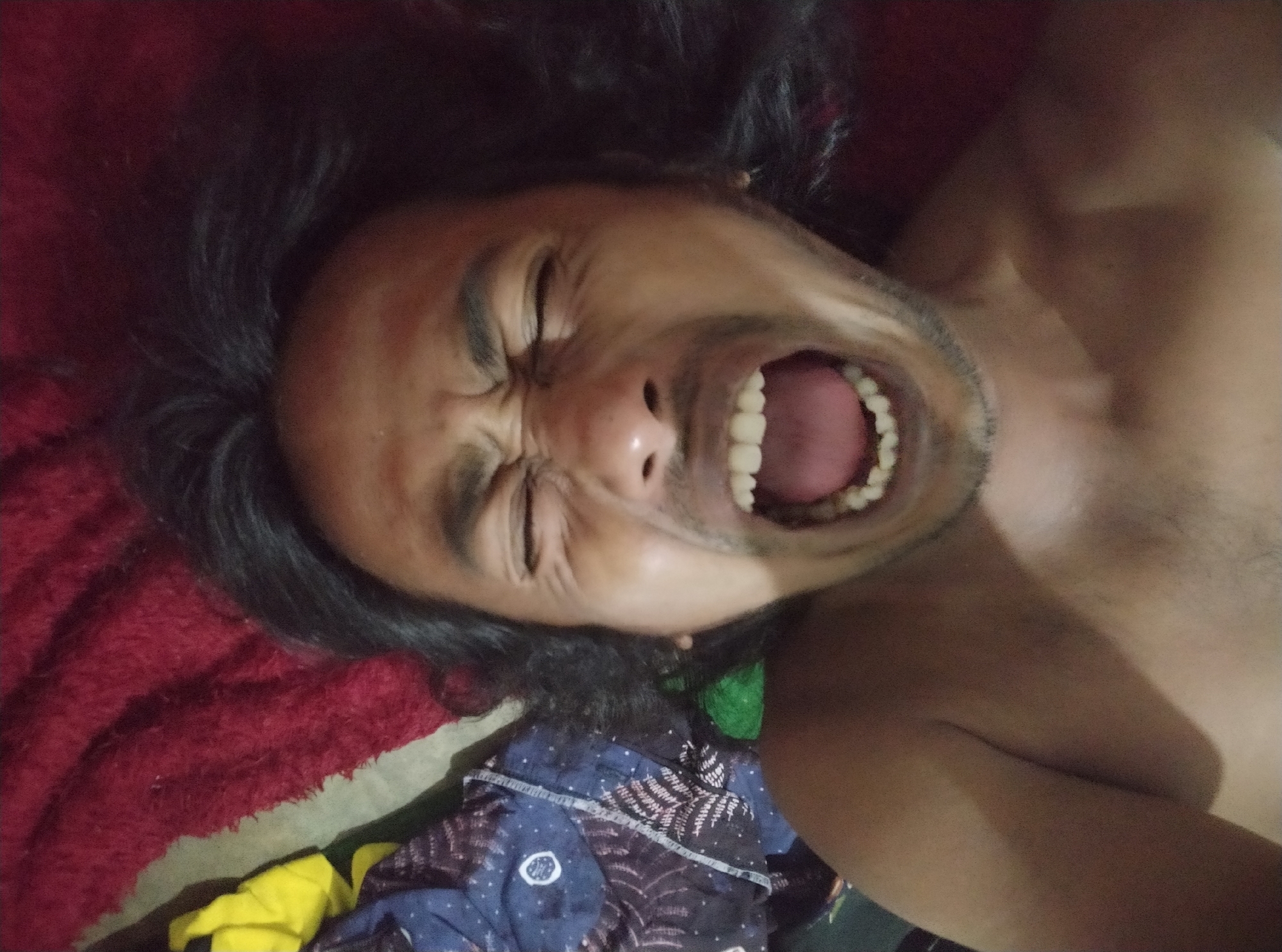 The image, taken in Sumurber, Indonesia, shows a person lying on their back on a red blanket. Their mouth is open wide, revealing teeth and tongue, and their eyes are squeezed shut, suggesting they are shouting or laughing intensely. The person has long, dark, messy hair that spreads out around their head. In the background, various fabrics are visible, including a blue patterned cloth and a bright yellow cloth. The scene appears to be indoors, possibly a bedroom.