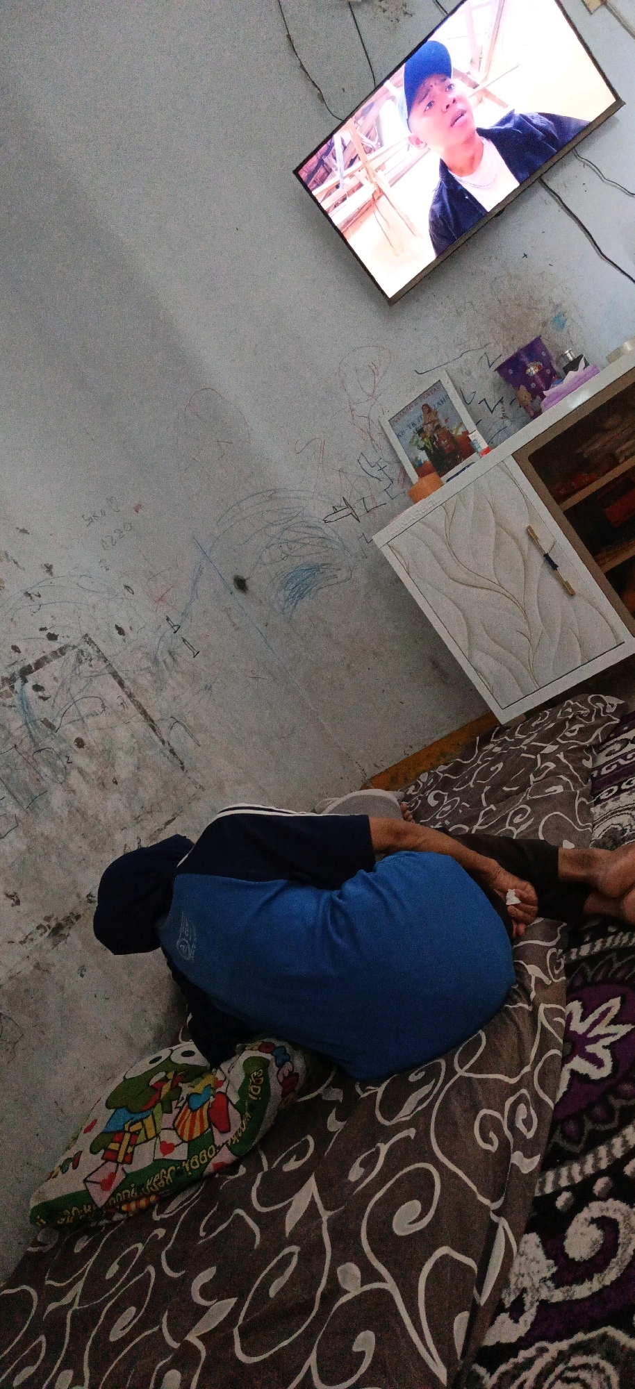 In a dimly lit room in Panjatan, Indonesia, a person lies on a bed covered with patterned blankets, facing away from the camera. They are wearing a blue shirt and dark pants, with their head tucked in. A colorful pillow with cartoon characters is placed near their head. On the wall, a flat-screen television displays a young man wearing a blue cap and a dark jacket. The wall itself is covered in various scribbled drawings, some appearing to be done with crayons. To the right, a white cabinet with decorative carvings stands near the wall. The overall impression is of a lived-in space, likely a bedroom.