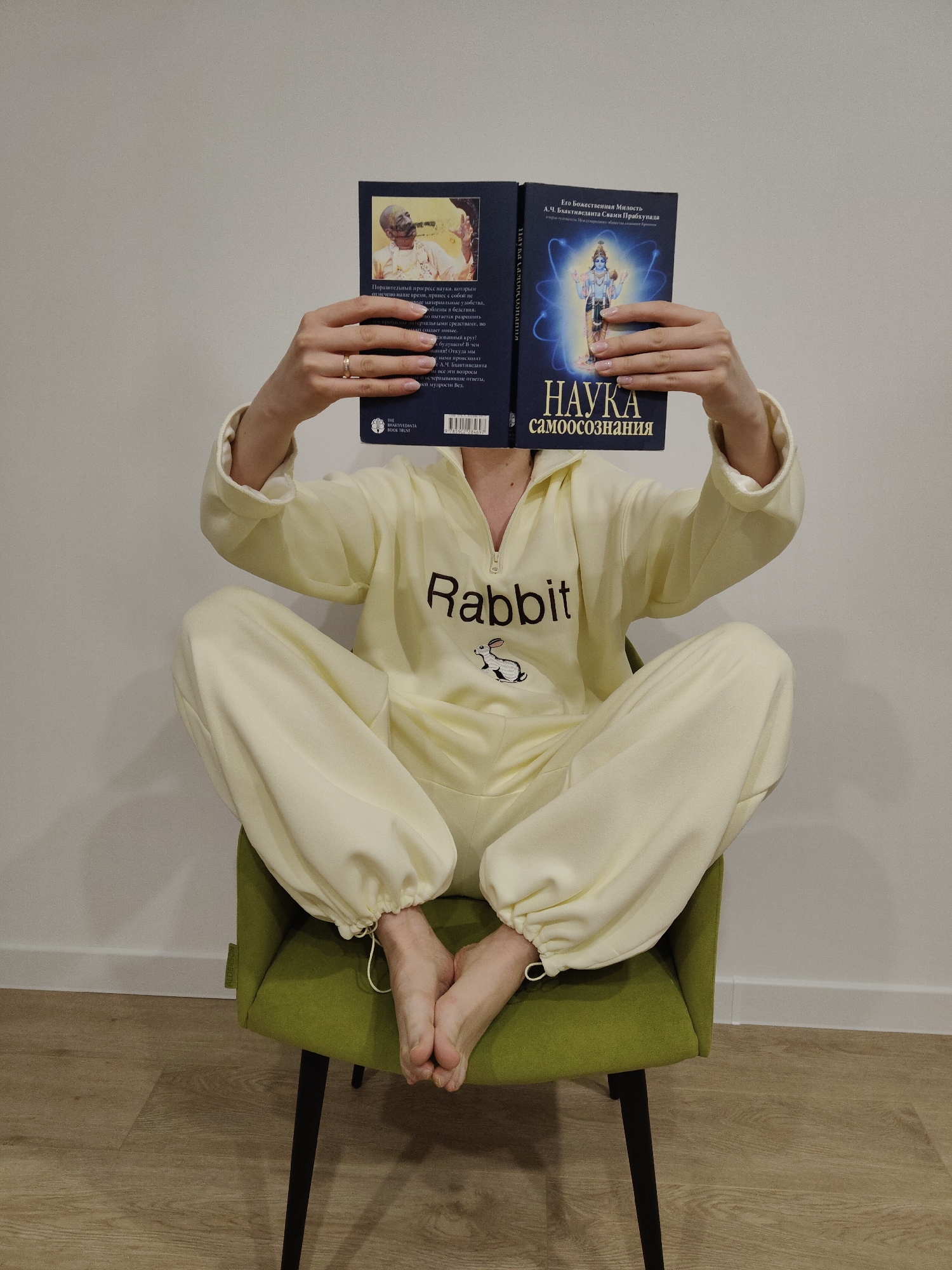 A person is seated on a muted green upholstered chair, holding an open book that obscures their face. They are wearing a light, pale yellow or cream-colored hooded tracksuit or onesie with a zipper detail and the word "Rabbit" embroidered on the chest, accompanied by a small rabbit illustration. Their legs are crossed, and bare feet are visible, with the tracksuit's pant legs gathered at the ankles by drawstrings. A ring is visible on the person's left hand, which holds the book.
The book has a dark blue cover with Cyrillic text. The visible front cover displays "НАУКА самоосознания" (Science of Self-Realization) and "Его Божественная Милость А.Ч. Бхактиведанта Свами Прабхупада" (His Divine Grace A.C. Bhaktivedanta Swami Prabhupada), featuring an illustration of a blue-skinned deity with a halo. The partially visible back cover includes the text "THE BHAKTIVEDANTA BOOK TRUST" and a barcode, along with Cyrillic paragraphs discussing the progress of science, problems, and philosophical questions. An image of a man is also visible on the back cover.
The setting is indoors, against a plain white wall with a white baseboard. The floor is made of light-colored wooden laminate. The scene suggests a calm moment of reading or contemplation.