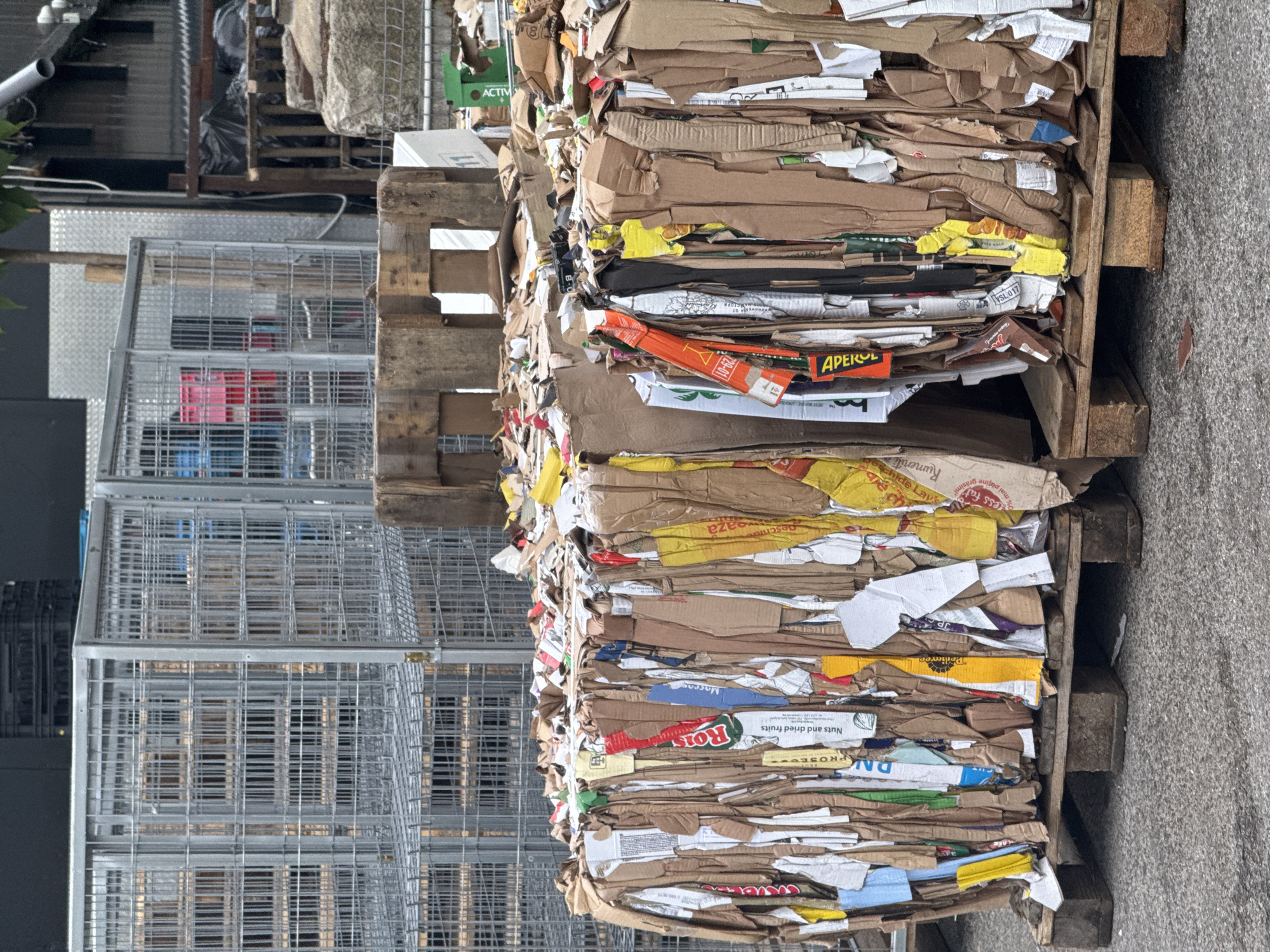 This is a high-angle, medium shot of a recycling facility in Sofia, Bulgaria. In the foreground, two large, tightly compressed bales of cardboard sit on wooden pallets. The bales are composed of various cardboard pieces, some with colorful labels and text. To the left, a metal cage filled with what appear to be empty plastic crates, some red and blue, is visible. In the background, more metal cages are stacked, along with a few large grey concrete blocks. The ground is paved with asphalt. The overall impression is one of an industrial recycling or waste management area.