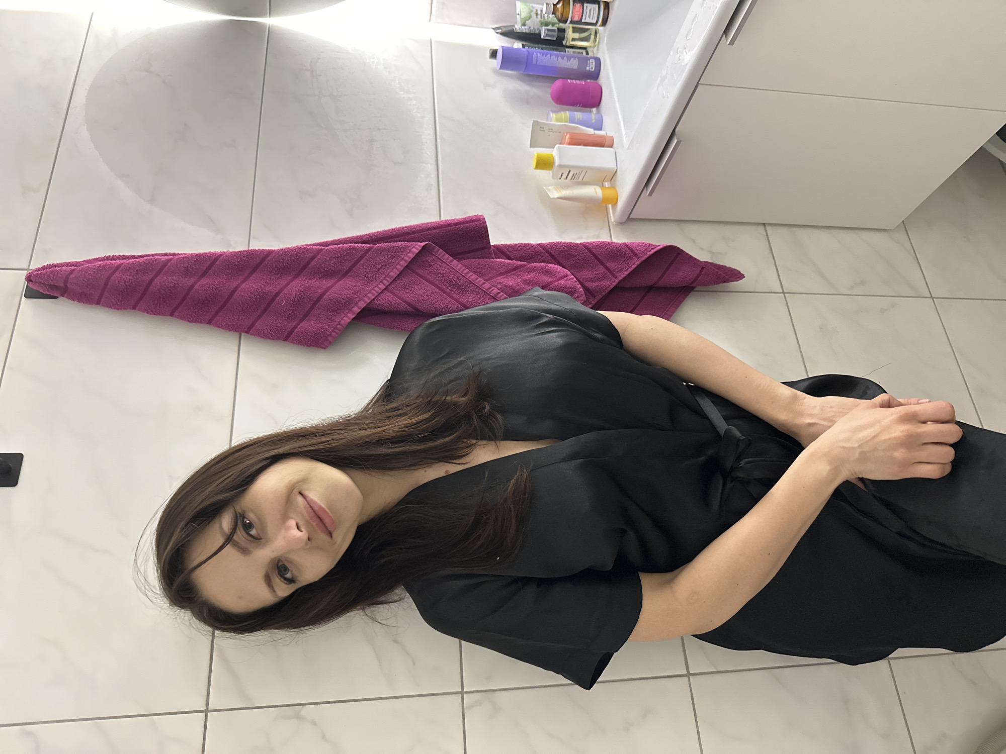 A woman in a black robe is lying on a tiled floor next to a pink towel in what appears to be a bathroom. A white vanity with toiletries sits in the background. The location is potentially Murino, Russia, but this cannot be confirmed from the image.