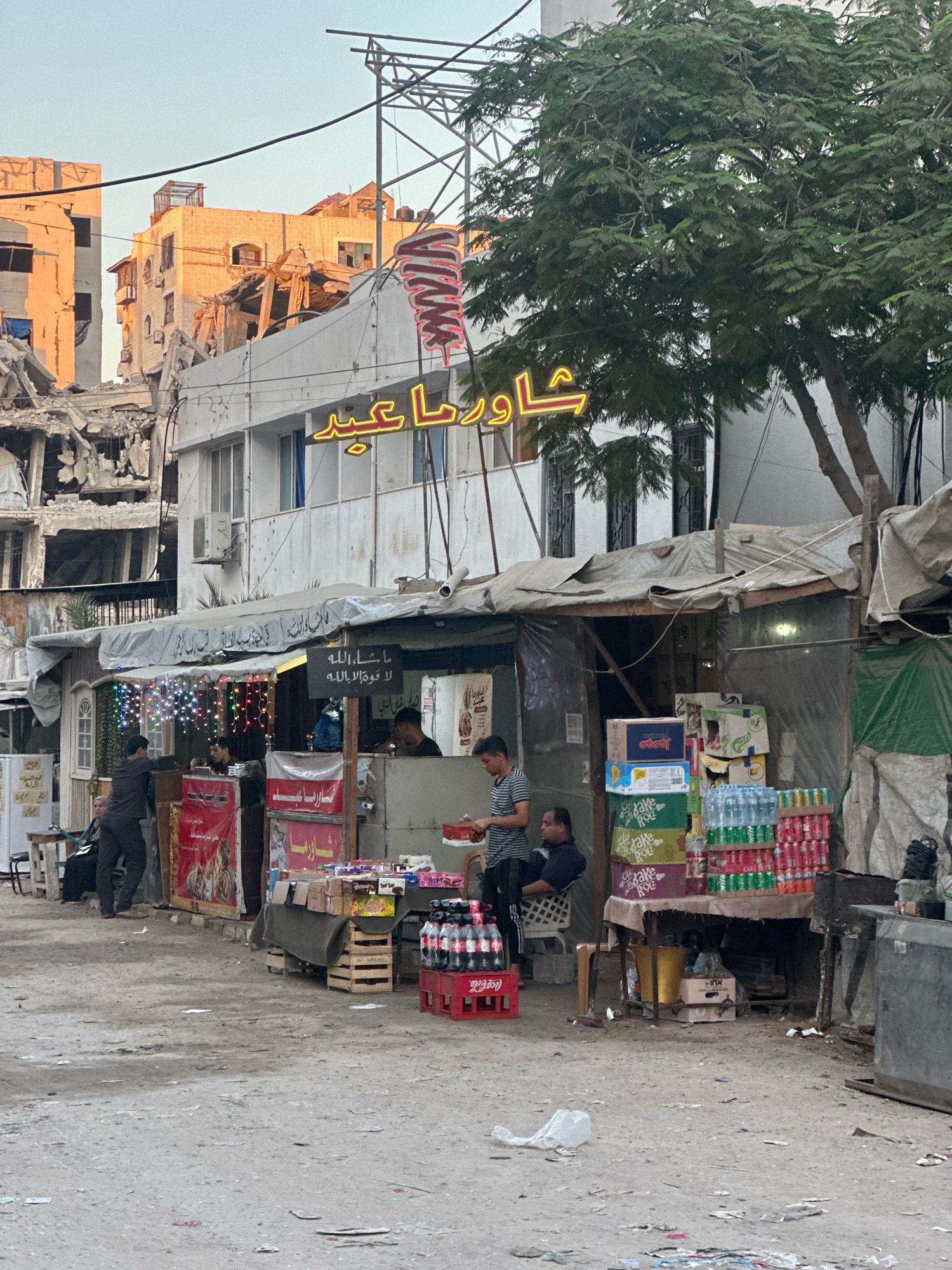The image depicts a street scene in the Gaza City, Palestinian Territories, characterized by a damaged building and a small food stall. The background shows a partially destroyed building with visible structural damage and debris, indicating the aftermath of conflict. In the foreground, a makeshift food stall is set up under a canopy. The stall displays various items, including drinks in bottles and cans, food boxes, and what appears to be advertising banners. Several individuals are present around the stall; some are standing and interacting with the stall owner or customers, while others are seated. The ground is dusty and scattered with debris and litter. A prominent neon sign with Arabic lettering is visible above the stall. The overall atmosphere suggests a scene of resilience and everyday life continuing amidst difficult circumstances.