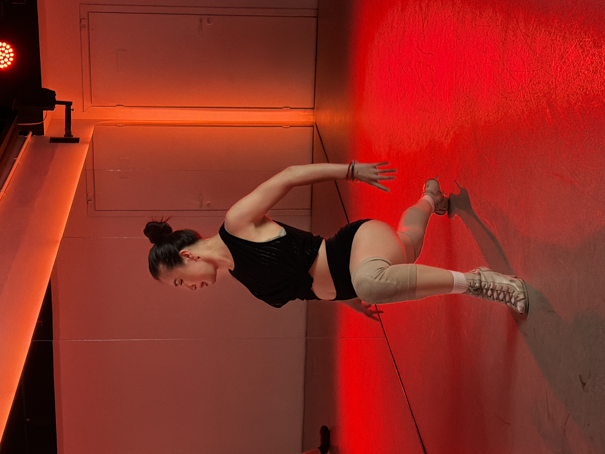 A young woman wearing a black top, black shorts, a knee brace, and sneakers is in a dance studio. The studio appears to be indoors, featuring red lighting. Her hair is in a bun and she is in the middle of a dance move with one arm extended and a slightly arched back. The location is Moscow, Russia, but the studio specifics cannot be derived from the image.