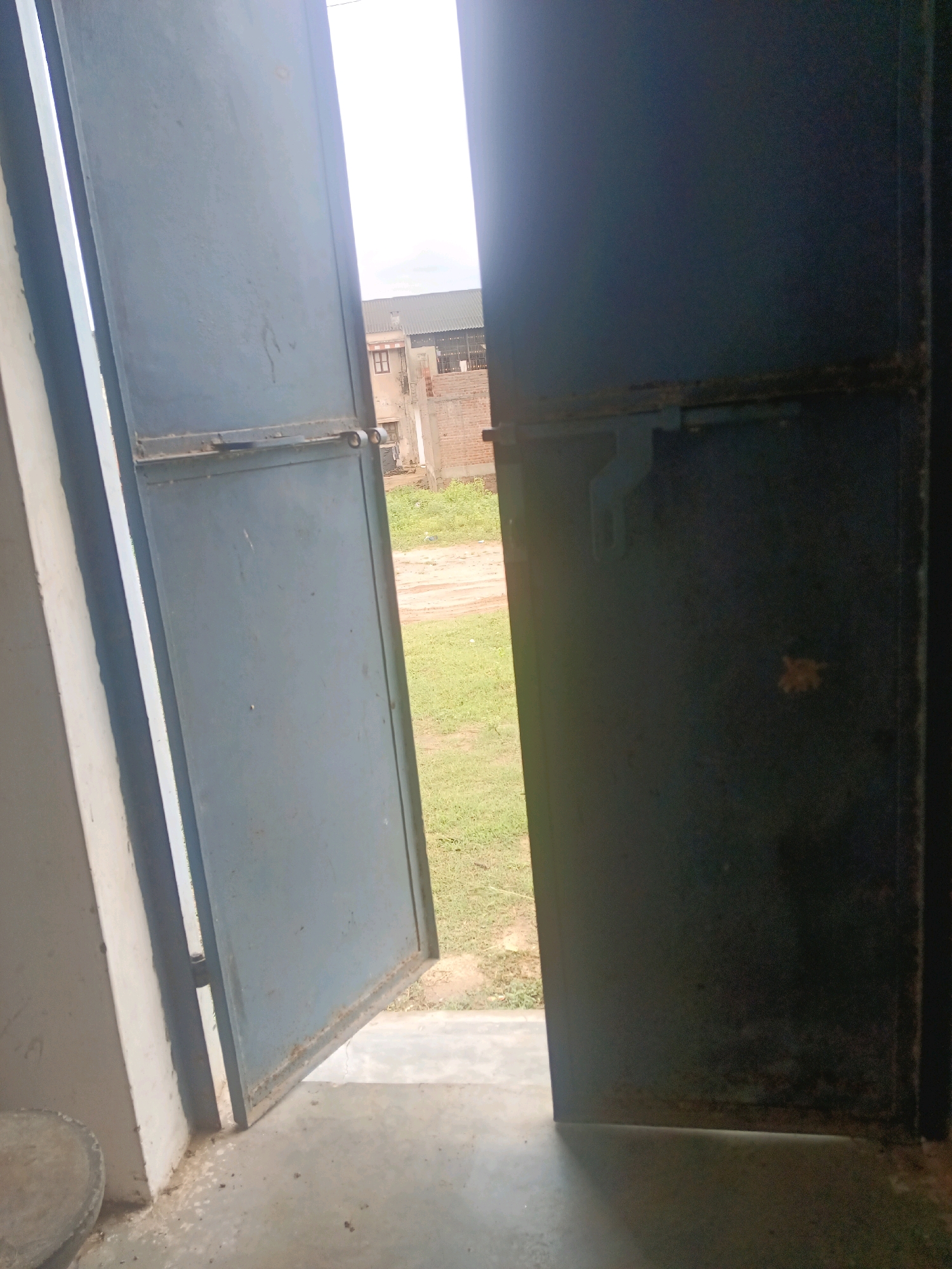 The image depicts an outdoor scene looking through an open, dark blue metal gate. The gate appears to be ajar, revealing a glimpse of the exterior. To the left, a portion of a light-colored wall is visible. The ground inside the gate is a concrete surface, with some debris. Beyond the gate, there is a patch of green grass and a dirt path. In the background, several brick buildings are visible, suggesting an urban or semi-urban setting. One building has a grey metal roof. The sky is bright and appears to be overcast. The image is taken from a low angle, looking slightly upwards. There are no people or animals visible in the frame. The scene appears to be located in Tokariya, India, as indicated by the prompt.