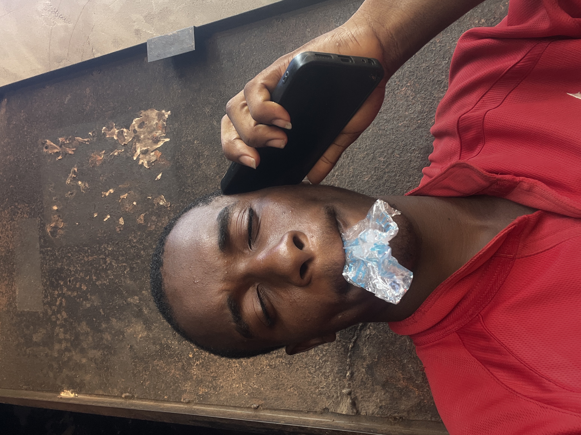 A man lies sideways on a dark, gritty surface, possibly metal, with peeling paint. He wears a red shirt and has a small plastic bag filled with liquid in his mouth. He is holding a black smartphone to his forehead. The location is unknown, but the question asks to consider the location as Onitsha, Nigeria.