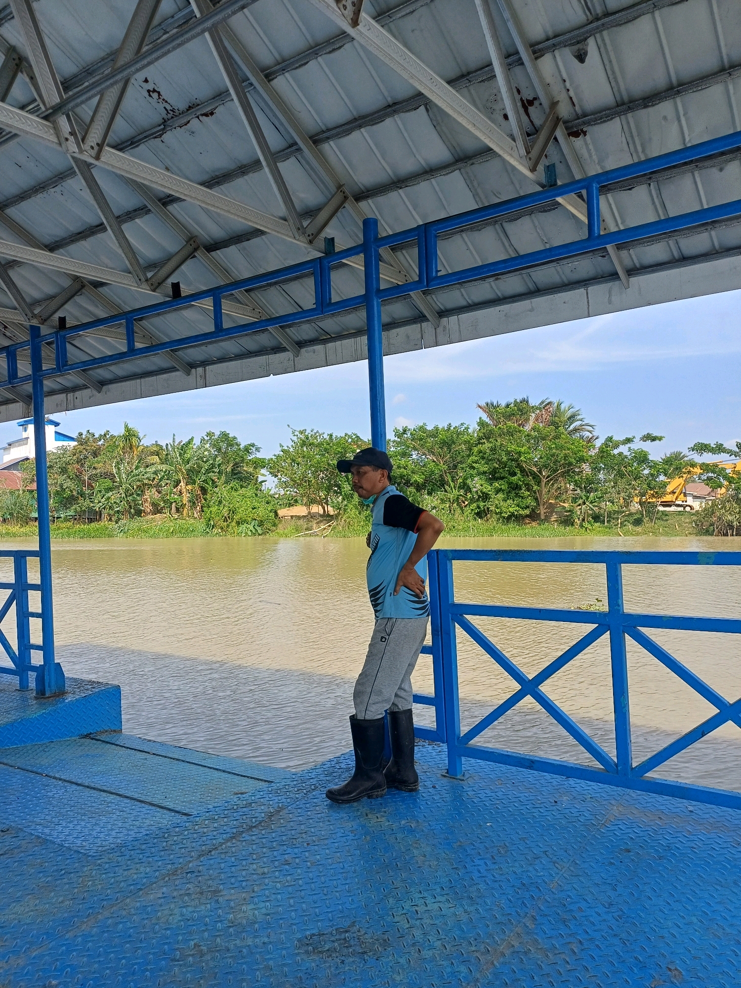 A man stands on a blue metal platform beneath a silver corrugated metal roof supported by white metal trusses and blue metal railings. He is wearing a black cap, a blue and black t-shirt, grey pants, and black rubber boots. The man is facing to his right, with his left hand on his hip. The platform is next to a wide, muddy brown river. Across the river is a densely vegetated bank with various trees, including banana trees, and some small buildings and structures. In the background, a yellow construction vehicle is partially visible among the trees. The sky is clear and blue. The location is identified as Pasir Belengkong, Indonesia.