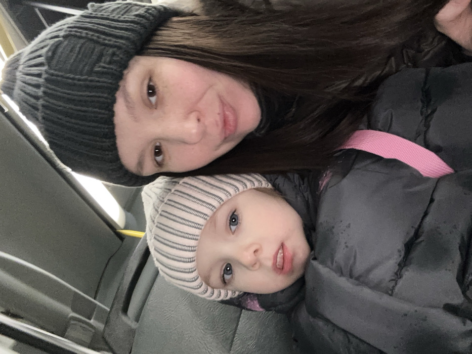 A woman and a child are seated, likely indoors. The woman wears a dark gray knit hat and has long dark brown hair. The child, with striking blue eyes, sports a striped gray and white knit hat and is bundled in a black puffer jacket with a pink strap visible. Both are looking at the camera, close together, in what appears to be a selfie. The setting suggests public transport, given the visible seat and interior paneling.