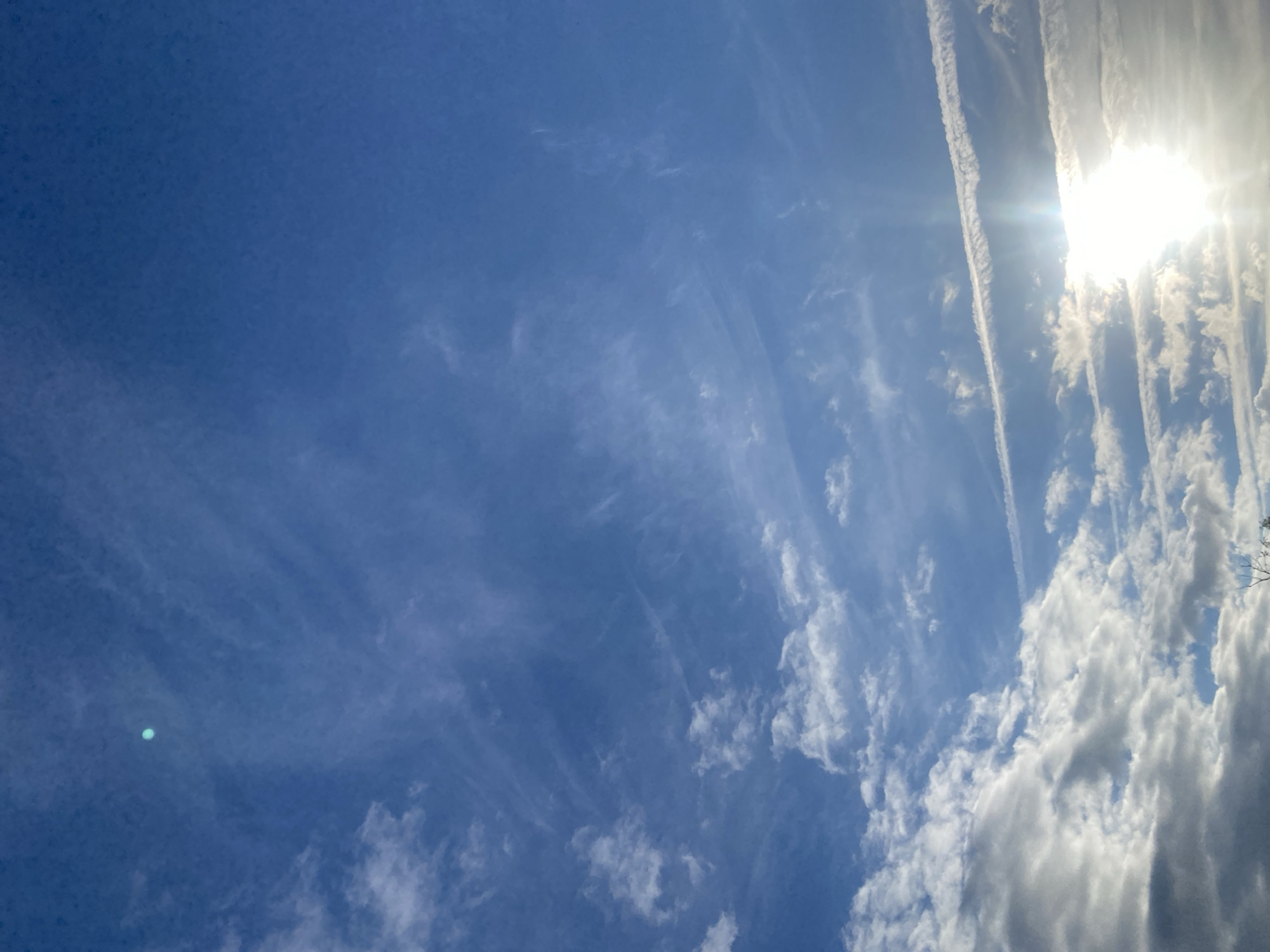 The image depicts a bright, clear sky with scattered clouds over what appears to be a rural landscape. In the upper left quadrant of the sky, a small, luminous object, possibly a lens flare or a distant balloon, is visible. The main body of the sky is a vibrant blue, streaked with wispy cirrus clouds. Towards the lower right, the sun is shining brightly, partially obscured by clouds and creating a lens flare effect. Several contrails from aircraft are also visible, forming linear patterns across the sky. The lower portion of the image shows the tops of trees against the bright sky, suggesting a natural or semi-natural environment. The location is identified as Байқоныс ауылдық округі, Kazakhstan.
