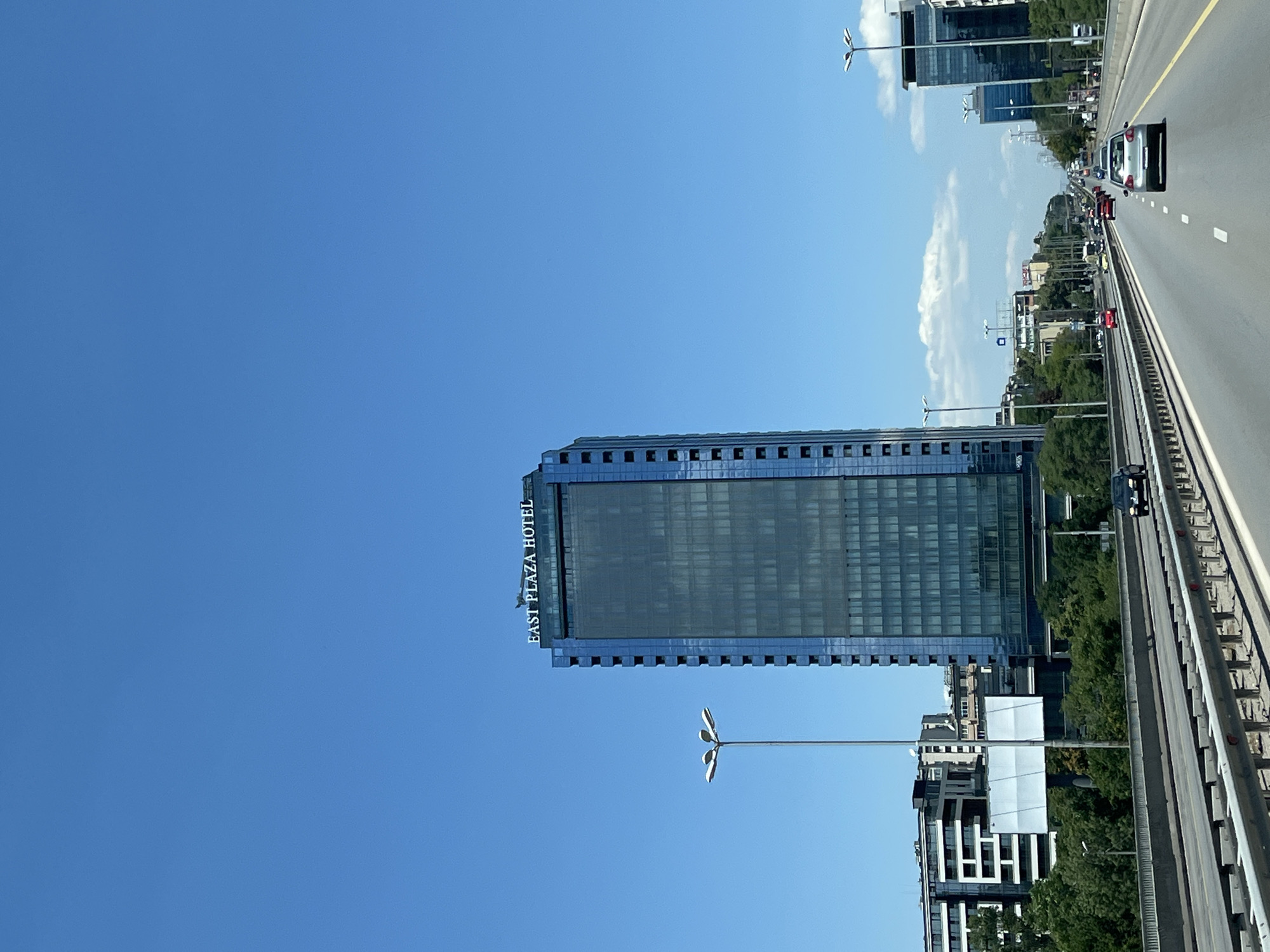 The image shows a multi-lane highway in a city under a clear blue sky. A tall, glass-facade skyscraper with "EAST PLAZA HOTEL" visible on top is the dominant structure on the left side of the road. Cars are visible on the highway, moving in the same direction. Other buildings and trees line the sides of the highway, suggesting an urban environment. There are streetlights along the road. The perspective is from a moving vehicle, looking forward.