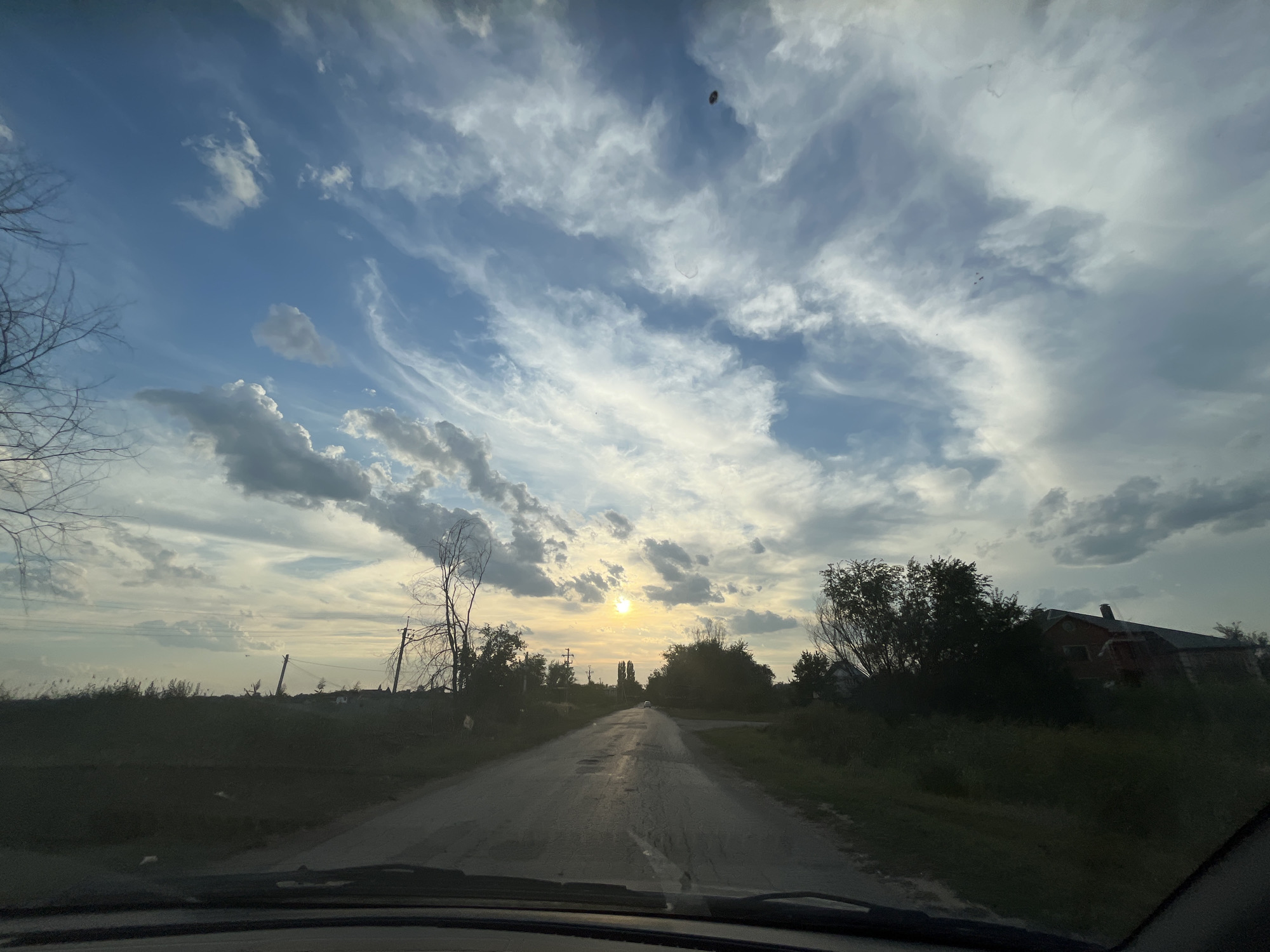 The image captures a rural road in Shumeyka, Russia, during what appears to be sunset or sunrise. The sky is filled with dramatic clouds, with streaks of white and grey against a blue backdrop. The sun is low on the horizon, casting a warm glow that reflects on the wet asphalt road. The road itself is in a state of disrepair, with visible patches and cracks.
To the sides of the road, there are dark, silhouetted trees and vegetation, some of which are bare and appear dead, while others are full of leaves. In the distance, the outlines of buildings, possibly houses, are visible amidst the greenery. Power lines and utility poles are also present along the roadside, suggesting a sparsely populated area with infrastructure.
The perspective of the image suggests it was taken from inside a vehicle, as the lower part of the frame shows the dashboard and windshield wiper. There are no visible people in the frame. The overall atmosphere is serene and slightly melancholic, characteristic of a quiet rural setting at twilight.