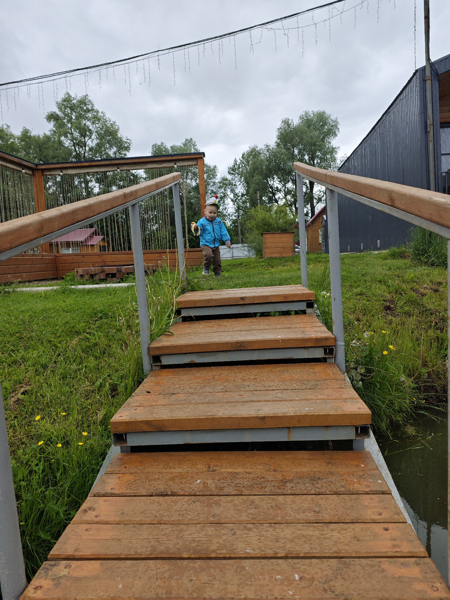 This image captures a low-angle perspective of a young child descending a grassy slope towards a wooden staircase, set in Malyi Bukor, Russia, under an overcast sky. The child, wearing a blue jacket, brown trousers, and a multi-colored hat, holds a yellow, banana-shaped object in their right hand. The wooden staircase, constructed with horizontal planks and supported by gray metal frames, extends from the foreground upwards and slightly to the right, featuring wooden handrails atop metal posts on both sides.
To the left of the staircase, a lush green grassy area is visible, dotted with small yellow dandelions. To the right, another patch of grass borders a dark body of water or a ditch, partially visible at the bottom right. In the middle ground, behind the child, there is a wooden fence or structure, and a small building with a red roof is partially obscured by trees. Further to the right in the background, a modern building with a dark gray, corrugated metal exterior is present. Above the scene, a string of decorative, icicle-like lights is suspended horizontally across the frame against the cloudy sky. Various green trees with dense foliage are scattered across the background.