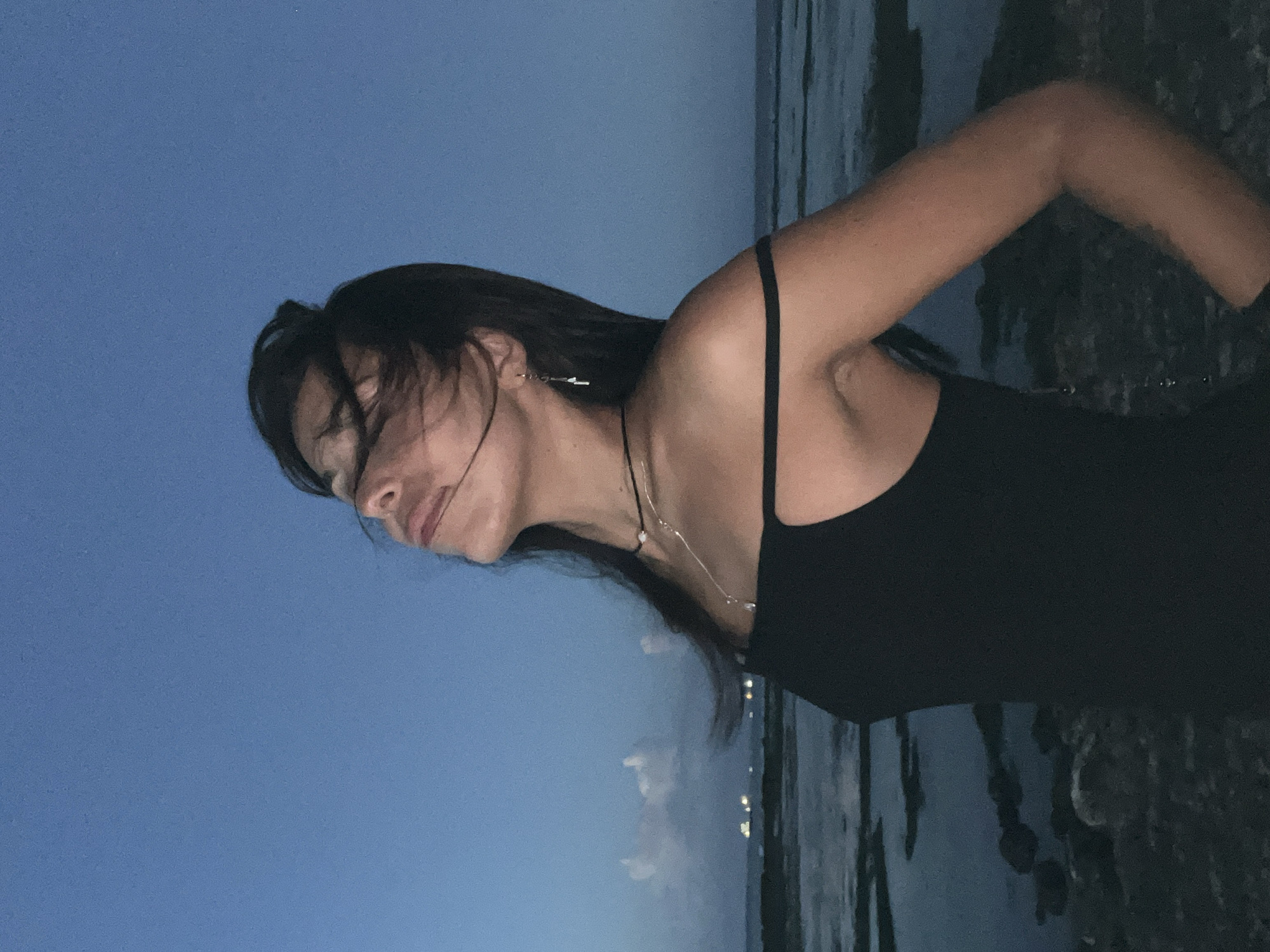 A person, likely female, is standing in what appears to be a coastal area in Kuta Selatan, Indonesia, at dusk or night. The individual is wearing a black top, with a necklace. The background consists of a dark blue sky transitioning to a horizon line with possible water and rocky formations. Lights from a distant area can be seen on the horizon. The individual's hair is windswept.