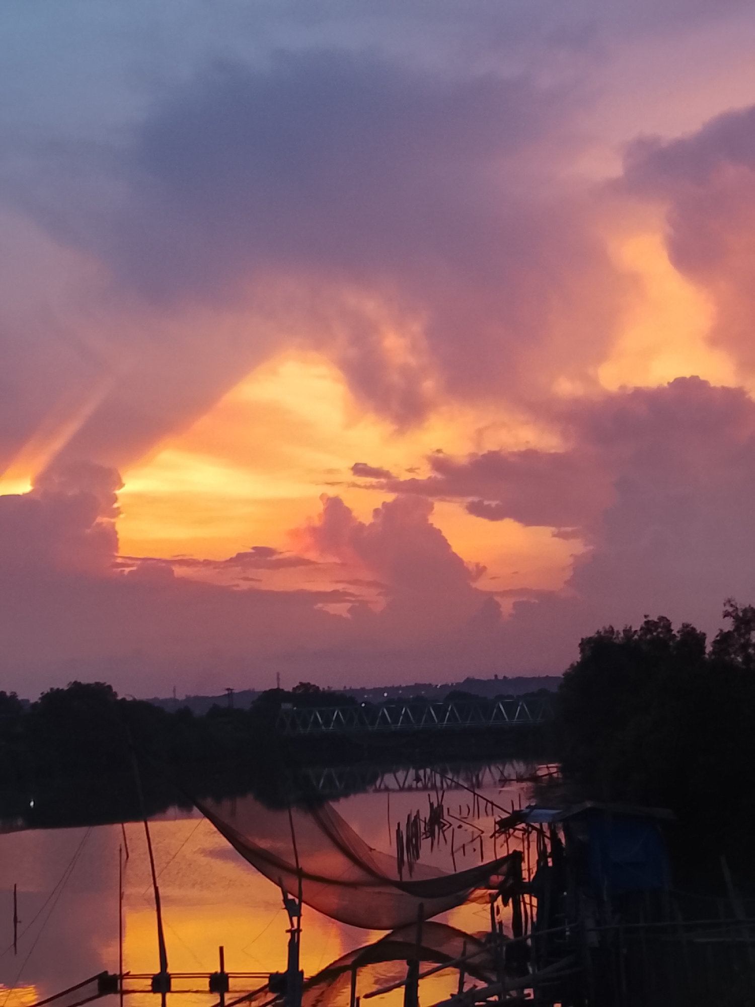 The image depicts a vibrant sunset or sunrise over a body of water, likely a river or estuary, in Lhokseumawe, Indonesia. The sky is ablaze with spectacular colors, ranging from intense oranges and yellows near the horizon to soft pinks, purples, and blues higher up. Dramatic, voluminous clouds are illuminated from below, creating a dynamic interplay of light and shadow. On the left, distinct crepuscular rays, or "god rays," pierce through the clouds, emphasizing the powerful light source hidden behind them.
The water below acts as a mirror, reflecting the brilliant hues of the sky, casting warm, shimmering colors across the surface. In the foreground and mid-ground, several traditional fishing structures are prominent. These appear to be fixed lift nets, large scoop nets suspended by wooden poles and frames over the water. On the right, a small, makeshift dwelling or platform structure, partially covered with a blue tarp, is visible amidst the fishing gear.
The banks of the water body are lined with dark silhouettes of dense trees and vegetation. In the middle ground, spanning the water, a distinctive metal truss bridge with a zigzagging pattern is clearly visible against the brighter horizon, suggesting developed infrastructure in the area. The distant shoreline also features a dark line of trees and possibly some indistinct buildings. The scene evokes a sense of tranquil beauty, capturing a moment of natural grandeur as day transitions to night or vice versa, set against a backdrop of local fishing activity. No visible text is present in the image.