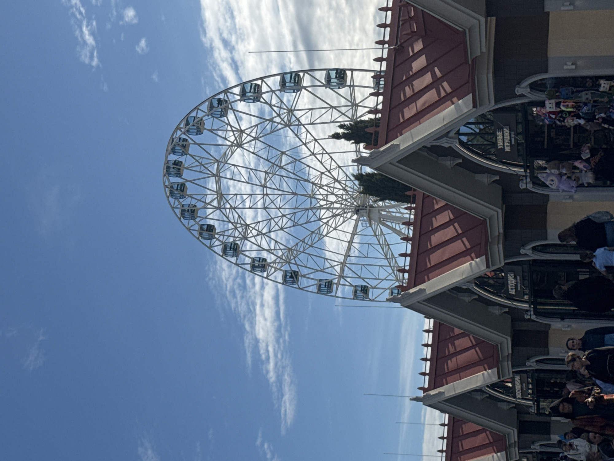 The image features a Ferris wheel against a blue sky with wispy clouds. The Ferris wheel has a white frame and enclosed gondolas. Adjacent to the Ferris wheel is a building with a red roof and arched doorways. People are gathered near the building's entrances. A sign with Russian text is visible. The location is the city of Sirius, Russia.