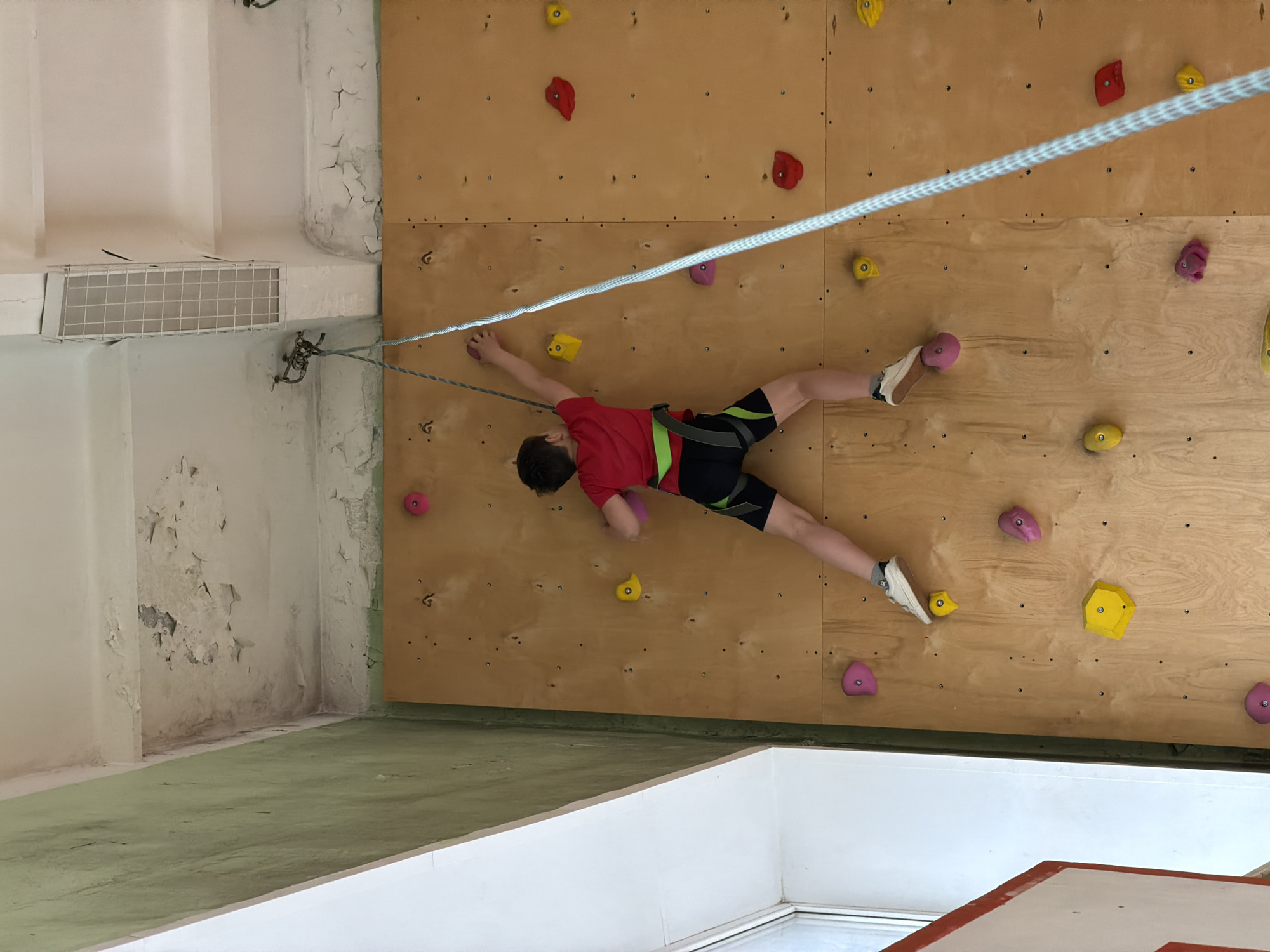 An indoor climbing facility in Izhevsk, Russia, is depicted from an elevated perspective looking down onto a climbing wall. A young person, possibly a boy, wearing a red t-shirt, black shorts with green trim, and a safety harness, is actively climbing horizontally across a wooden bouldering wall. The climber is mid-movement, stretching their left arm to grasp a yellow hold while their right hand is on a pink hold. Their right foot is planted on a pink hold, and the left foot is extended, poised to find the next foothold.
A white and grey climbing rope runs diagonally across the frame, attached to the climber's harness and extending to an anchor point on a white wall to the left. This anchor point is a metal carabiner setup. The climbing wall itself is made of light-colored wooden panels, fitted with numerous colorful climbing holds in shades of red, yellow, and pink.
The surrounding environment shows signs of age; the white walls and ceiling structures to the left and bottom exhibit considerable peeling paint and cracks. Below the climbing wall, a green floor area is visible, which appears to be a mat or painted concrete. A white metal grate is affixed to the wall near the rope's anchor point. In the bottom right, a white and red structure, possibly part of another climbing element or a safety mat, is visible. The lighting is bright and even, suggesting an indoor, artificially lit environment. No text is visible in the image.