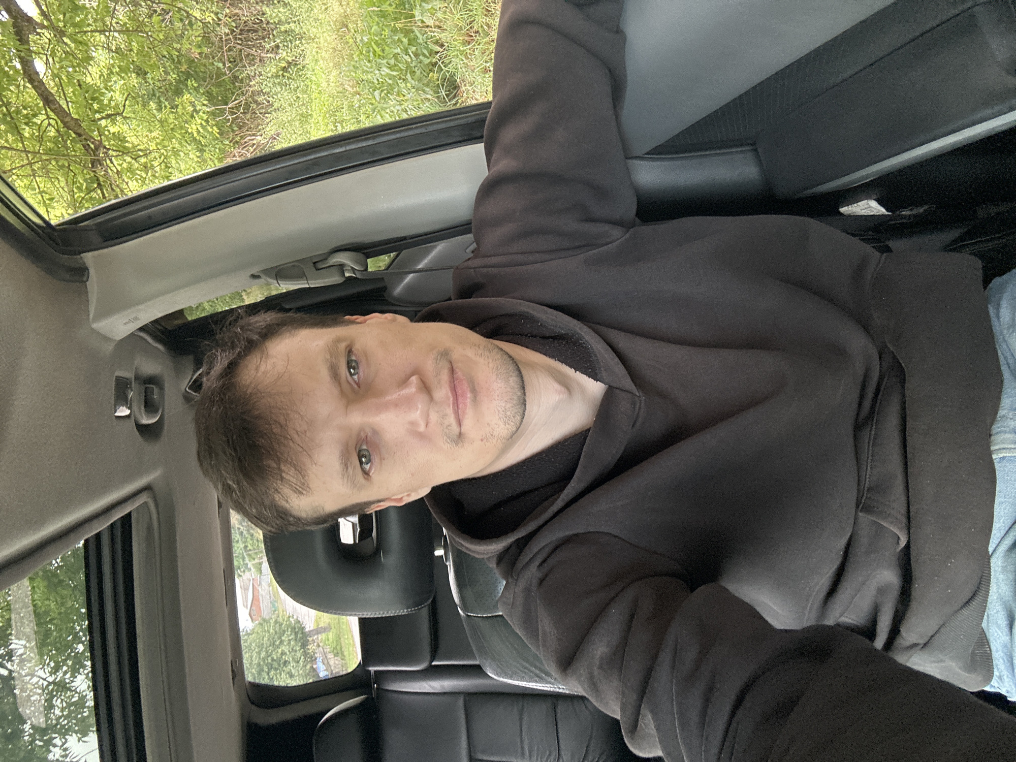 A medium shot shows a man with light brown hair and blue eyes, wearing a dark brown hooded sweatshirt and blue jeans. He is sitting in the driver's seat of a car. The car's interior is visible, including black leather seats and a sunroof. Outside the car, trees and greenery are visible, suggesting a rural or suburban setting. The image appears to have been taken in Makiv, Ukraine.