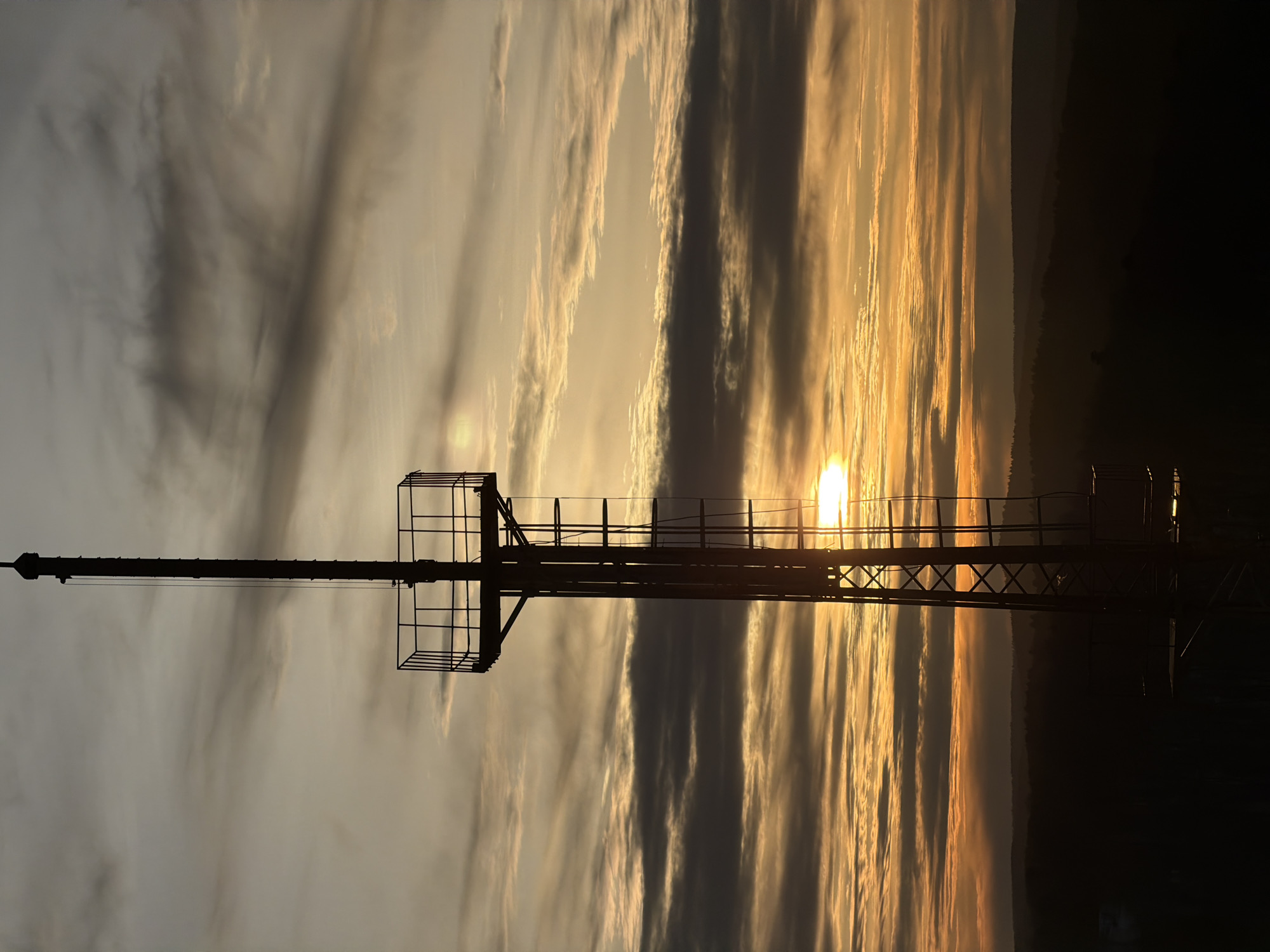 The image depicts a silhouette of a tower, likely a radio or communication tower, against a backdrop of a sunset. The sky is filled with striated clouds, colored with orange and yellow hues as the sun dips below the horizon. The tower is constructed of metal lattice and has platforms for maintenance or equipment. The surrounding area appears to be forested. While the exact location within Krasnoyarsk, Russia cannot be determined from the image alone, the presence of a tower and surrounding landscape suggest a location on the outskirts of the city or in a parkland area.