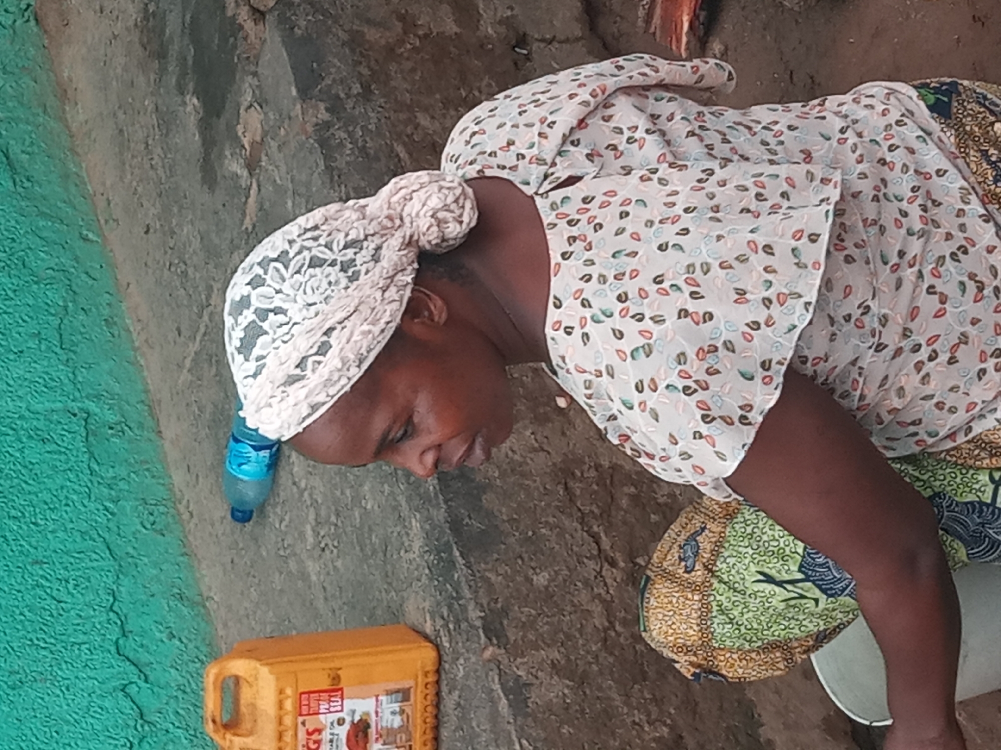 A woman is sitting on the ground outdoors in Sabon Tasha, Nigeria. She has dark skin and is wearing a light-colored headscarf with a lace pattern, a white floral shirt with short sleeves, and a patterned skirt in shades of green, yellow, and blue. She is looking down towards what appears to be a white bucket or container. To her left are a plastic bottle of water and a yellow container. The ground is rough and appears to be concrete or stone. A green painted wall is visible on the far left of the image.