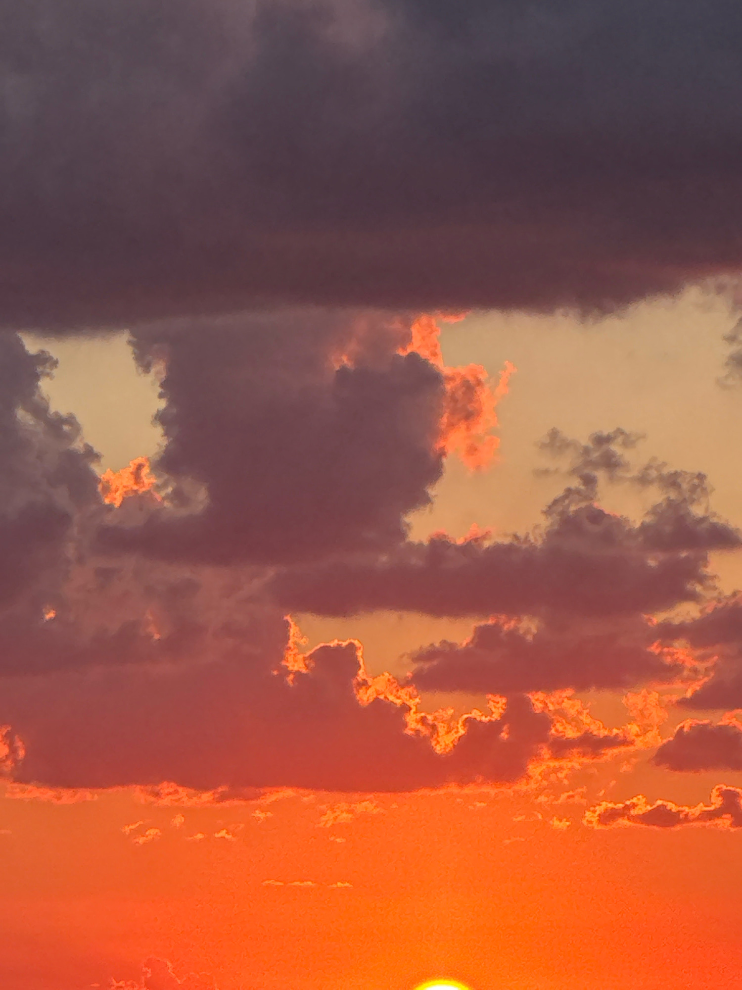 A sunset in Isola delle Femmine, Italy, features a vibrant sky filled with clouds in shades of orange, red, and purple. The bottom of the image is dominated by a brilliant orange glow, characteristic of a sunset. A sliver of the sun is visible at the very bottom, peeking through the horizon. The clouds are illuminated by the setting sun, creating dramatic contrasts and highlights. Some clouds appear dark and brooding, while others are edged with fiery orange light. The overall impression is one of a dynamic and colorful sky.